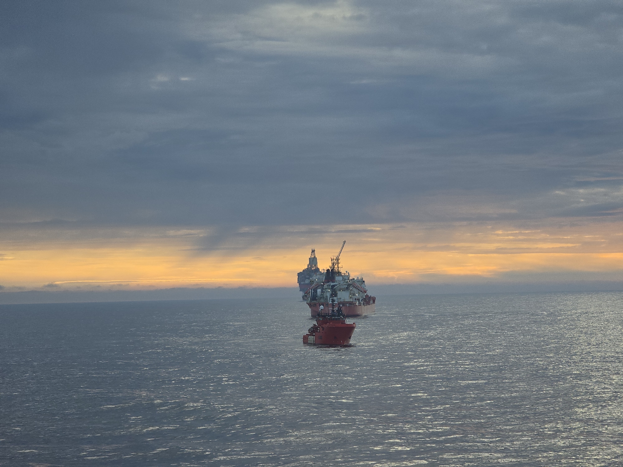 The image depicts three vessels on a body of water under a cloudy sky with a golden sunset glow on the horizon. The foreground vessel is a red supply ship, while the vessels behind appear to be oil rig platforms. The location is unknown.