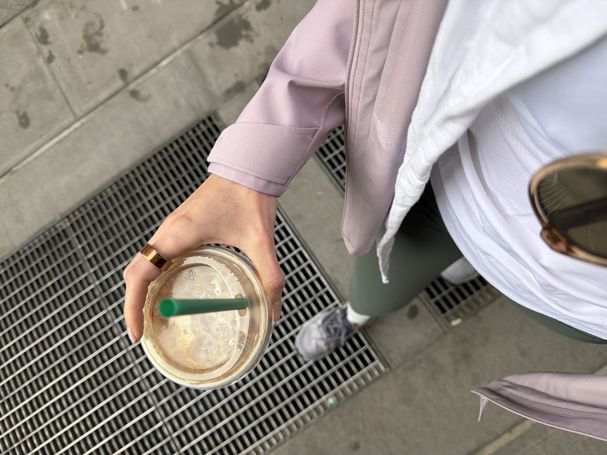 The image, captured from a high-angle perspective looking down, shows a person holding a clear plastic cup with a light brown beverage and a green straw. The person's right hand, adorned with a wide, gold-colored ring on the ring finger, holds the cup. The beverage appears to be an iced coffee or similar drink, with foam visible on top.
The individual is dressed in casual, layered attire. They are wearing a light purple jacket, possibly an athletic or windbreaker style, with visible seams and a slightly textured fabric. A white top is visible underneath the jacket, and dark green or grey leggings cover the legs. Part of a grey sneaker can be seen on the ground, suggesting the person is standing or walking. A pair of sunglasses, with a brown frame, is partially visible near the shoulder.
The setting is an urban outdoor environment, likely a sidewalk or street in New York, United States of America. The ground consists of large, light grey concrete slabs and a prominent metal grate, which covers a significant portion of the lower half of the frame.
Notable details include recycling symbols and material codes "PP", "5", "PA4", and "PET" molded onto the plastic lid of the cup. The image depicts a casual moment during daylight hours, but specific weather conditions or the exact time of day are not discernible. There are no strong emotional cues, and the focus is on the person's attire, the drink, and the urban ground elements.