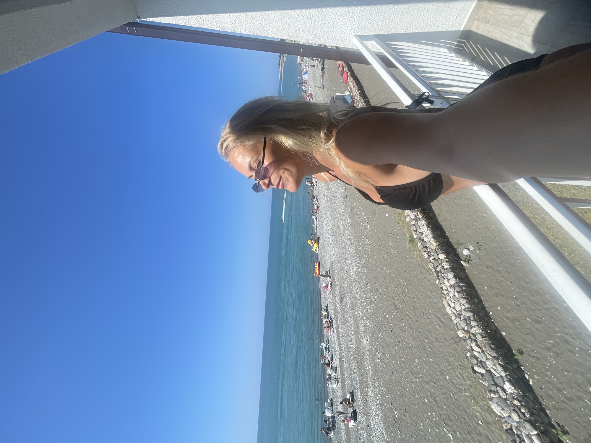 A blonde woman wearing a black bikini top and sunglasses is standing on a balcony overlooking a pebble beach and the sea. The sky is clear and blue. The beach is populated with people sunbathing on lounge chairs. The sea is a calm turquoise color. There are some inflatable toys in the water. The balcony has white railings and a white textured wall. The woman is looking out at the sea with her eyes closed, appearing to enjoy the sun. The photo is taken from a slightly low angle, looking up at the woman.