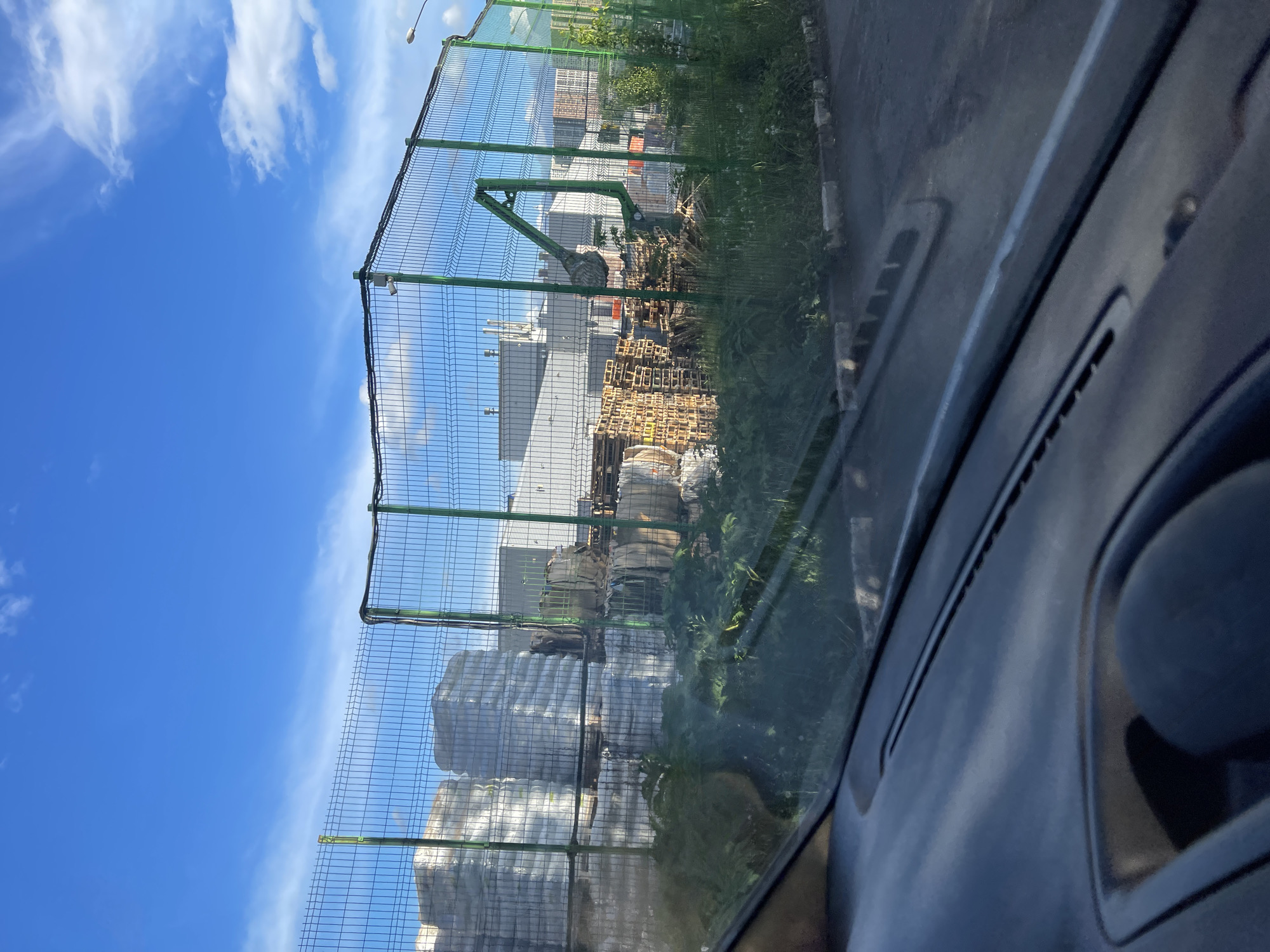 A view from inside a vehicle shows a fenced-off industrial area in Bittsa, Russia. Visible through a green wire mesh fence are stacks of white-wrapped materials, piles of wooden pallets, and industrial buildings. The sky above is blue with scattered clouds. Overgrown grass grows along the base of the fence. A portion of the vehicle's dashboard and window frame is visible on the right side of the image.