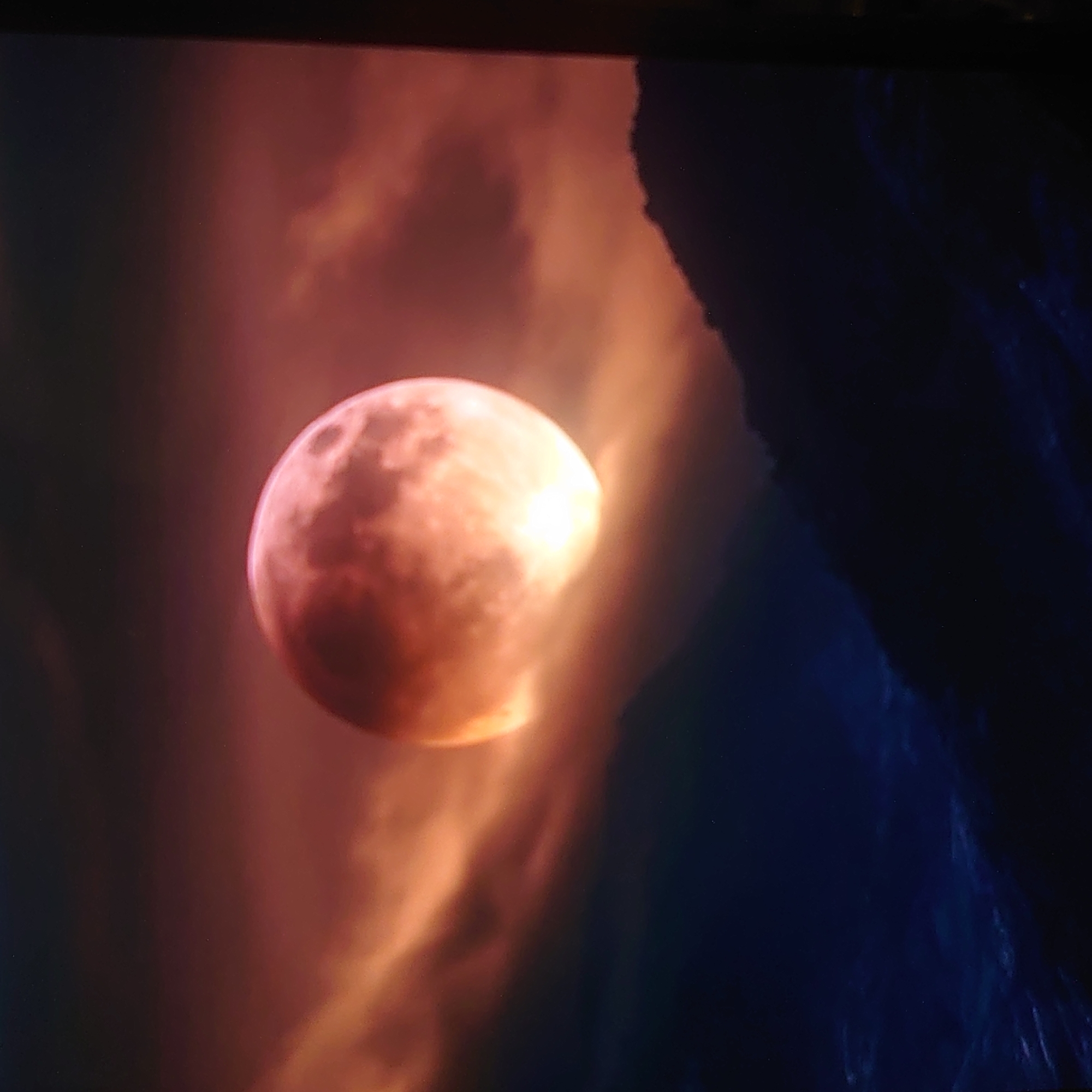 The image depicts a digitally rendered scene featuring a large, luminous, reddish-orange moon partially obscured by swirling, orange clouds. Below the moon, dark, silhouetted mountain ranges are visible against a deep blue, starry sky. The overall impression is one of a dramatic, otherworldly landscape. There is no discernible connection to the city of Belorechensk, Russia, nor are there any people, specific objects, or actions depicted.