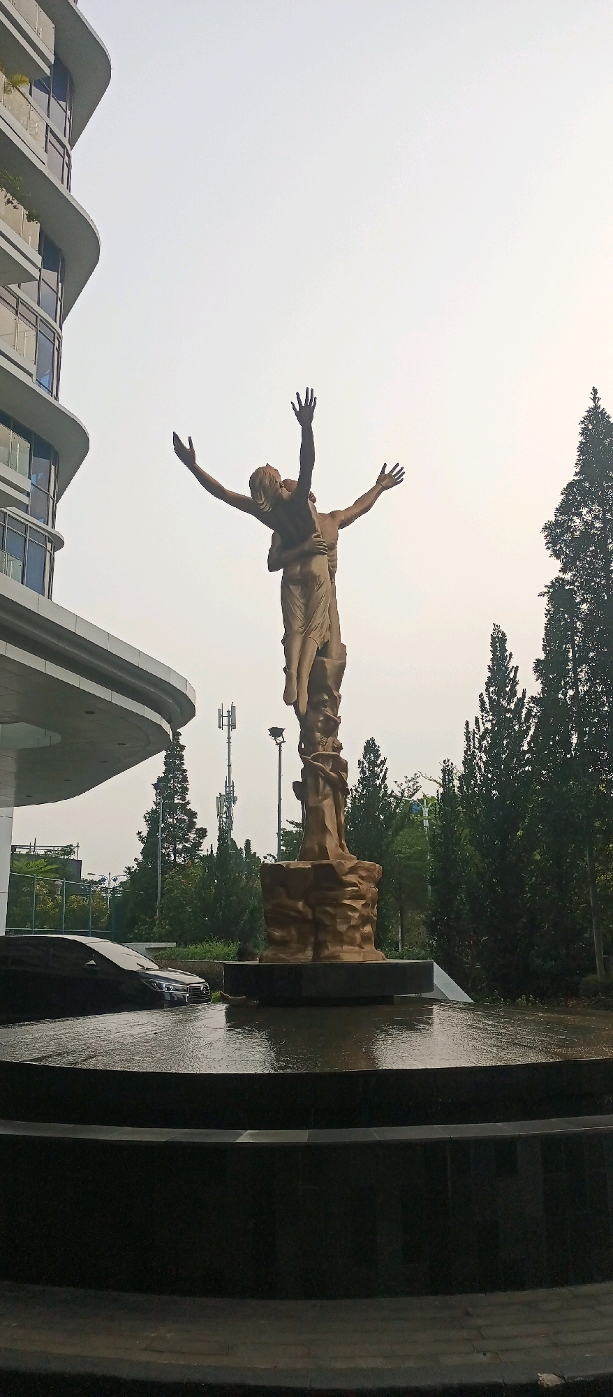 A bronze sculpture depicting two figures in an embrace stands in the foreground, with outstretched arms reaching towards a hazy sky. The figures are mounted on a rocky pedestal, which is surrounded by a shallow pool of water reflecting the overcast sky. To the left of the sculpture is a modern, curved building with balconies and large windows, typical of contemporary architecture. In the background, there are several tall trees and communication towers. A dark-colored car is partially submerged in the water to the left of the sculpture, suggesting a possible flooding event or an artistic water feature. The overall scene appears to be an outdoor public space, possibly a plaza or park adjacent to the building. This location is identified as Citra Towers in West Jakarta, Indonesia.