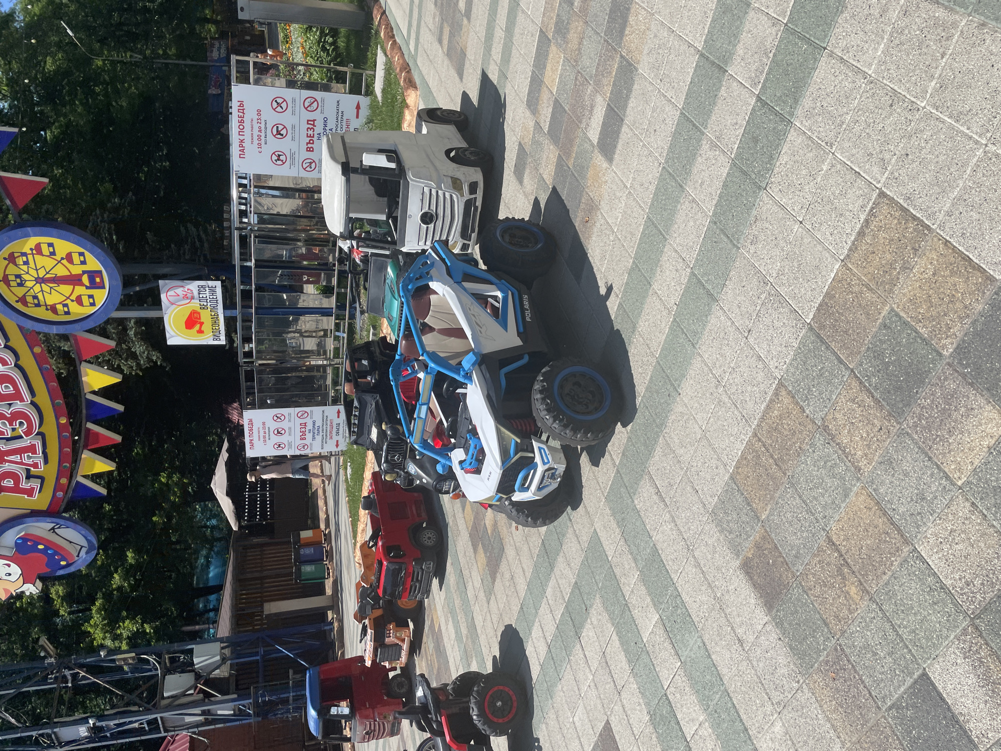 A sunny, outdoor scene in Stavropol, Russia, shows a variety of children's ride-on vehicles parked on a tiled walkway. In the foreground, a white and blue Polaris ATV is prominent, with large, knobby tires. Behind it, a white toy truck, resembling a semi-trailer, is parked. Further back, several other ride-on vehicles, including red trucks and a black off-road vehicle, are visible. A metal fence encloses part of the area, and a sign with Cyrillic text and pictograms is posted, indicating park rules and operating hours. Behind the fence, trees and some low structures are present. In the upper portion of the image, a colorful amusement park sign featuring a Ferris wheel and flags is visible, suggesting this is an entertainment area within the city. A person in a light-colored shirt and shorts walks away from the camera in the mid-ground. The overall impression is a recreational space for children.