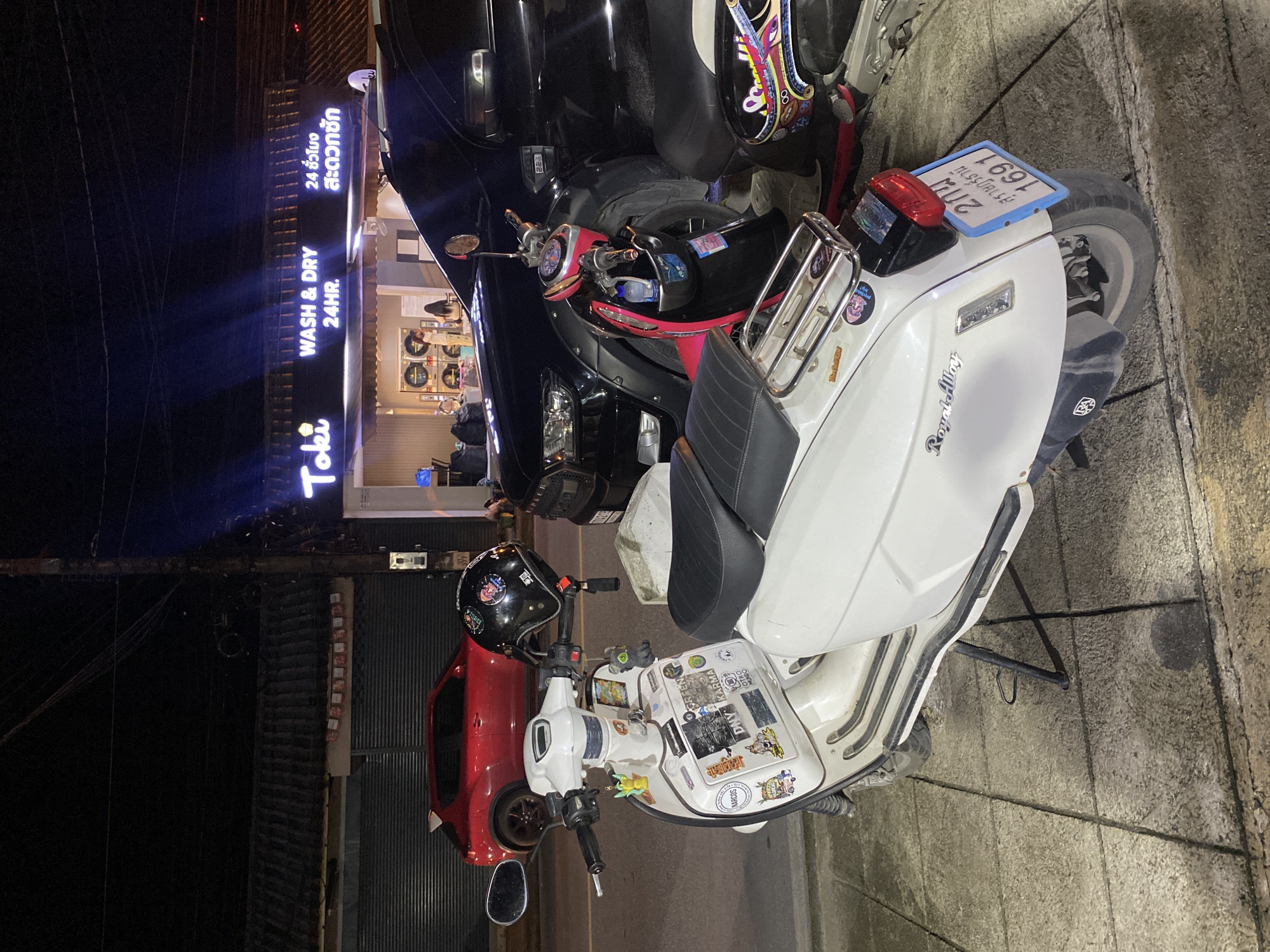 The image shows a street scene in Baan Chaweng Noi, Thailand, at night. Parked on the sidewalk are several scooters, including a white "Royal Alloy" scooter with numerous stickers and a license plate partially visible. A black helmet is placed on the scooter's handlebars. A red car is parked nearby. In the background, a laundromat named "Toki WASH & DRY 24HR." is visible, with a lit sign in both English and Thai. The street is paved, and the overall scene appears to be a typical urban environment in the area.