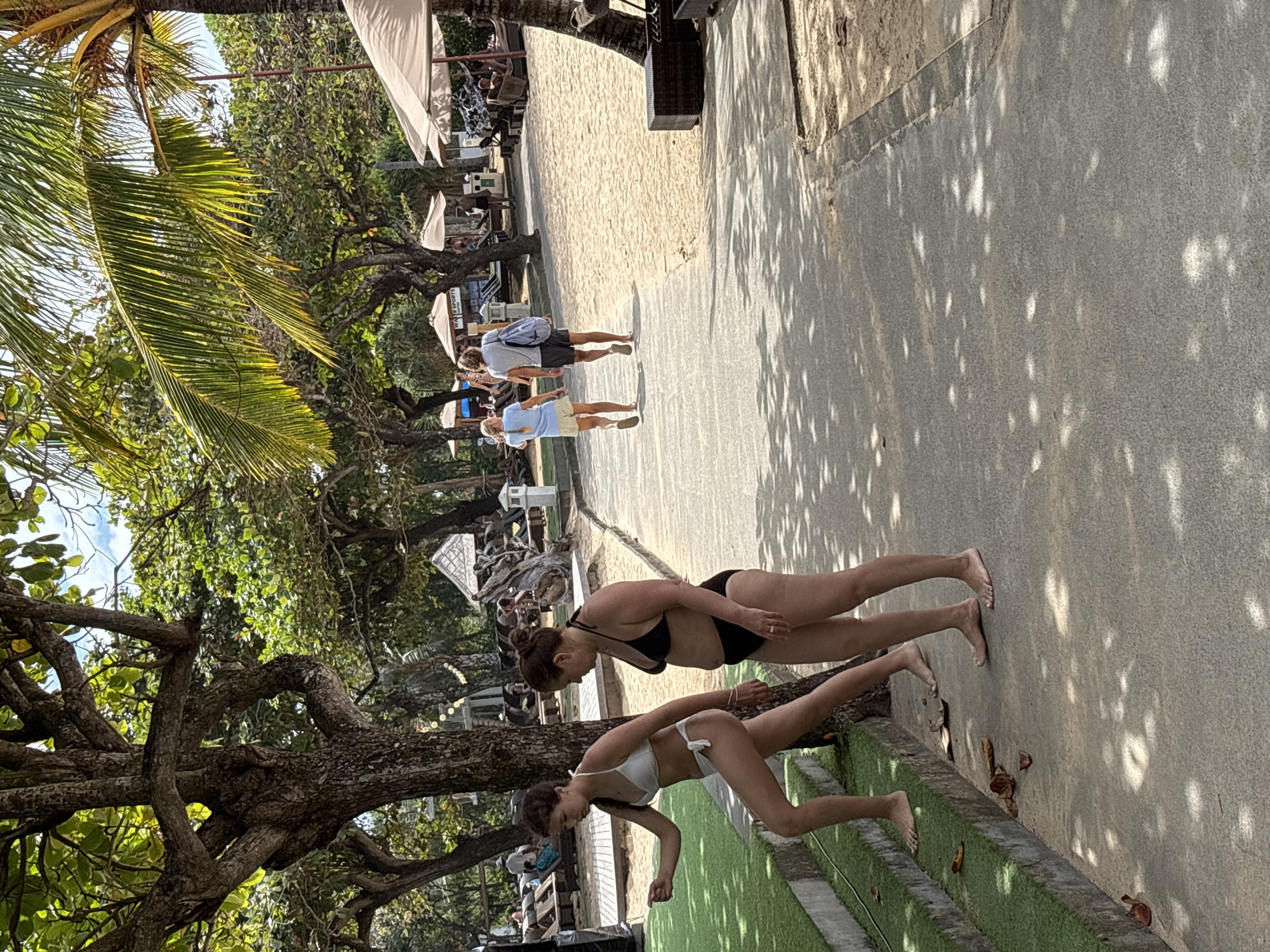 In Kuta Selatan, Indonesia, two women in bikinis are on a paved pathway near a sandy beach and lush trees. The woman on the left, with dark hair, wears a white bikini and is ascending a set of grass-covered stairs, gesturing with her left hand. The woman on the right, with dark hair pulled back, wears a black bikini and walks on the pathway. Further down the pathway, a couple walks away from the camera; the woman has blonde hair and wears a light blue t-shirt and shorts, while the man wears a grey t-shirt and dark shorts and carries a blue bag. Beach umbrellas and lounge chairs are visible on the sand, and palm trees and other foliage provide shade over the scene. The sky is bright with some clouds.