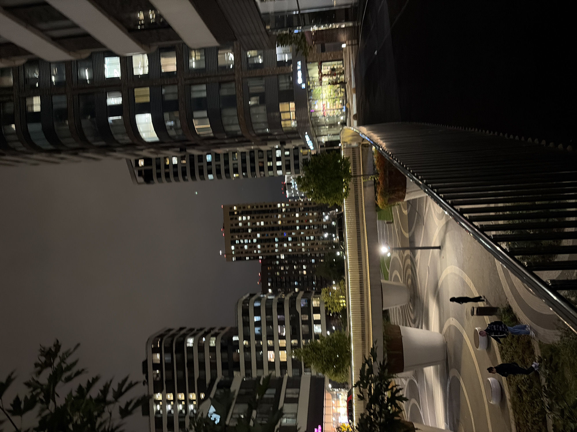 This is a nighttime photograph taken in Moscow, Russia, showing a modern residential complex. In the foreground, a dark metal railing runs diagonally across the frame, obscuring a portion of the view. Beyond the railing, a paved walkway is visible, with large, white planters containing greenery. Several people are walking on the walkway. In the midground, a pedestrian bridge curves across the scene, supported by large, cylindrical structures. Beyond the bridge, numerous high-rise apartment buildings are illuminated, their windows glowing with interior lights. Some of these buildings have a contemporary architectural style with curved facades. Streetlights and architectural lighting cast a warm glow on the surroundings. The sky is dark and overcast, typical of a city night. A few trees with dark foliage are also present in the scene.
