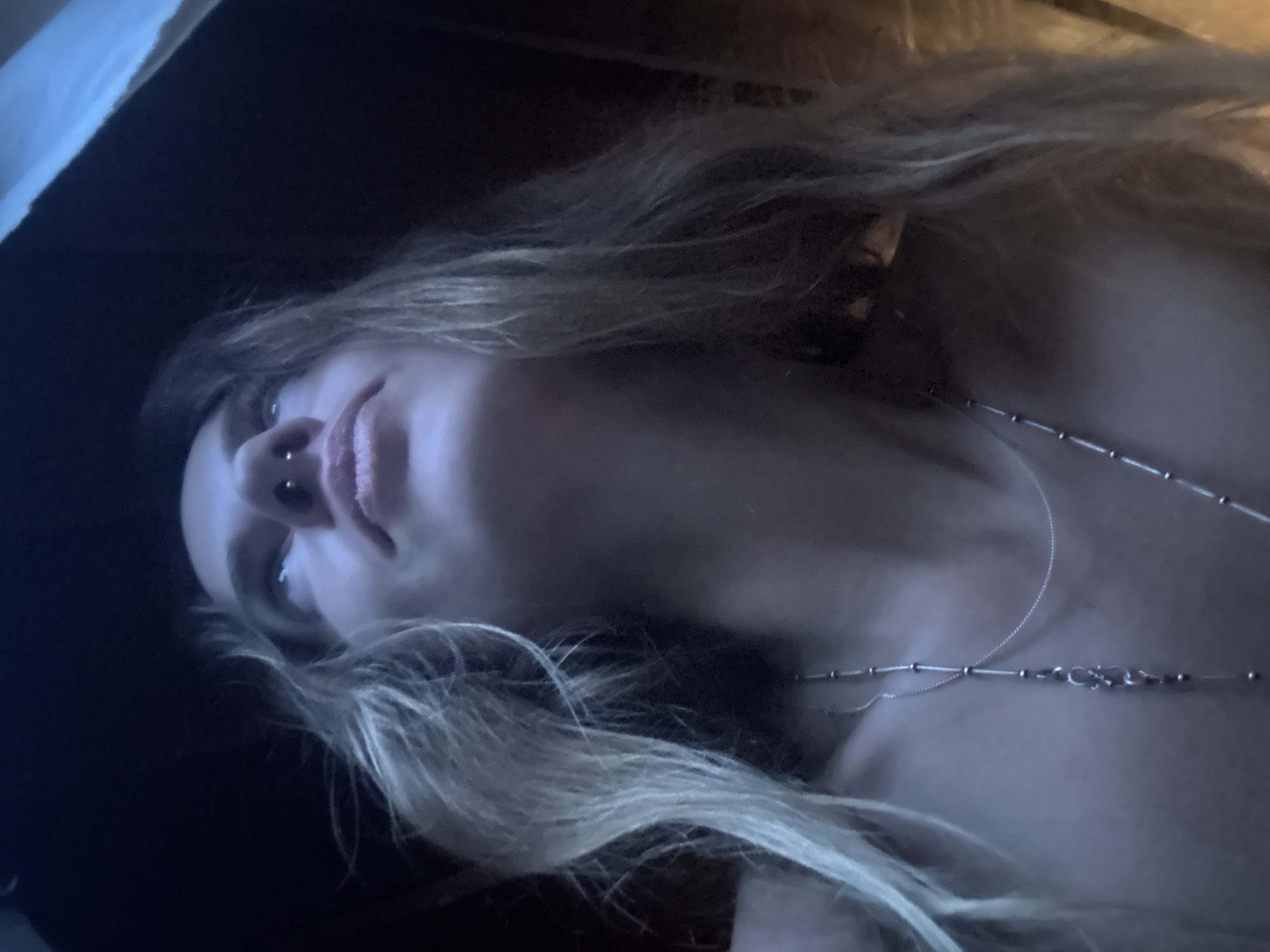 A low-angle, close-up shot shows the face of a person looking upward, framed by dark surroundings. The person has long, blonde hair, a nose ring, and is wearing two silver necklaces. The lighting is dim, with a blueish tint. The background is dark, suggesting an indoor or sheltered environment, possibly at night. The scene does not provide any specific information about the location within a city.