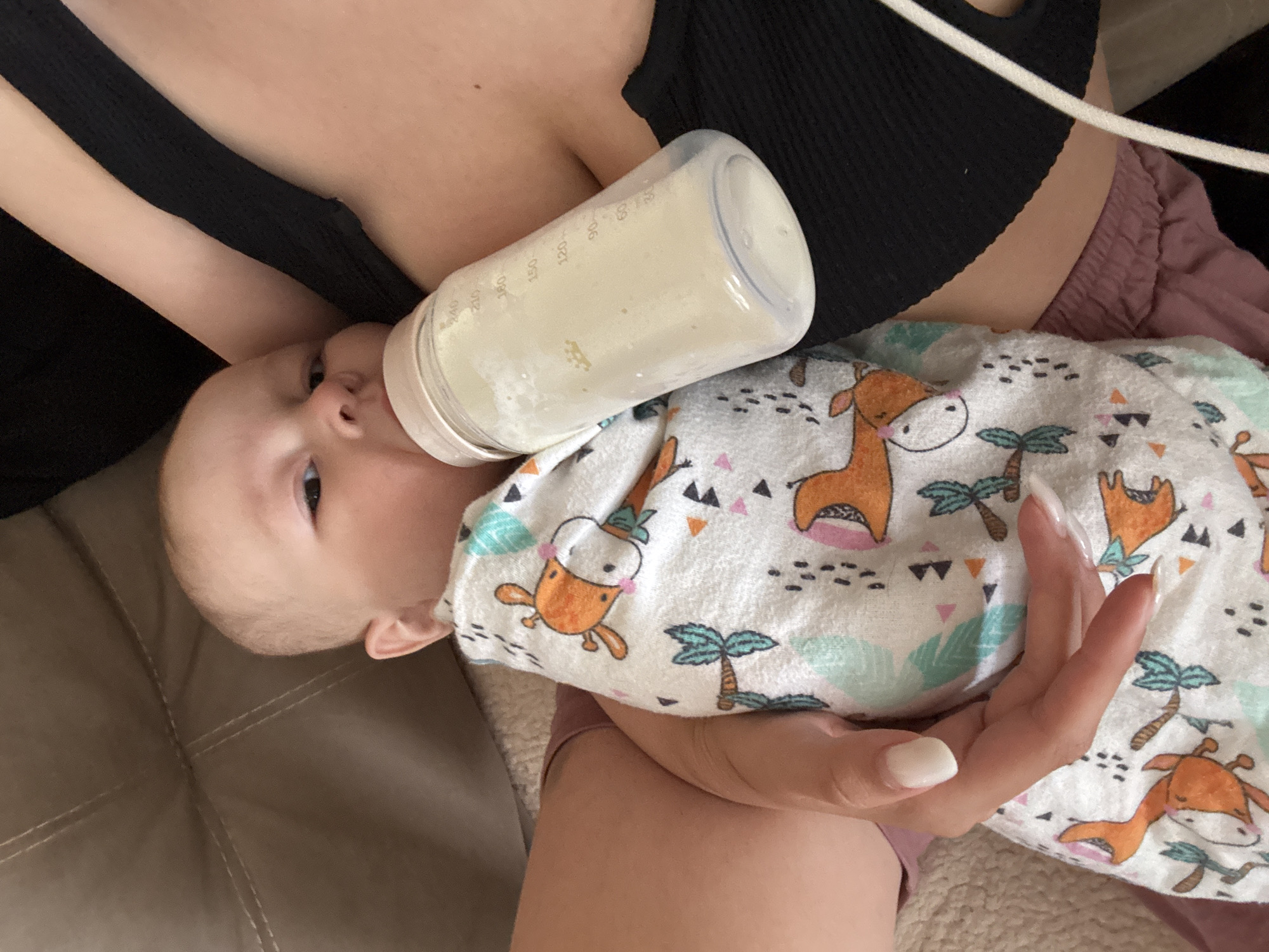 A baby is lying on a patterned blanket, being fed from a bottle held by an adult. The baby is looking at the camera with one eye visible. The adult is wearing a black top and pink shorts, and their hand with long white nails is visible. The scene is indoors on a brown couch. The location in Sochi, Russia, is not evident from this image.
