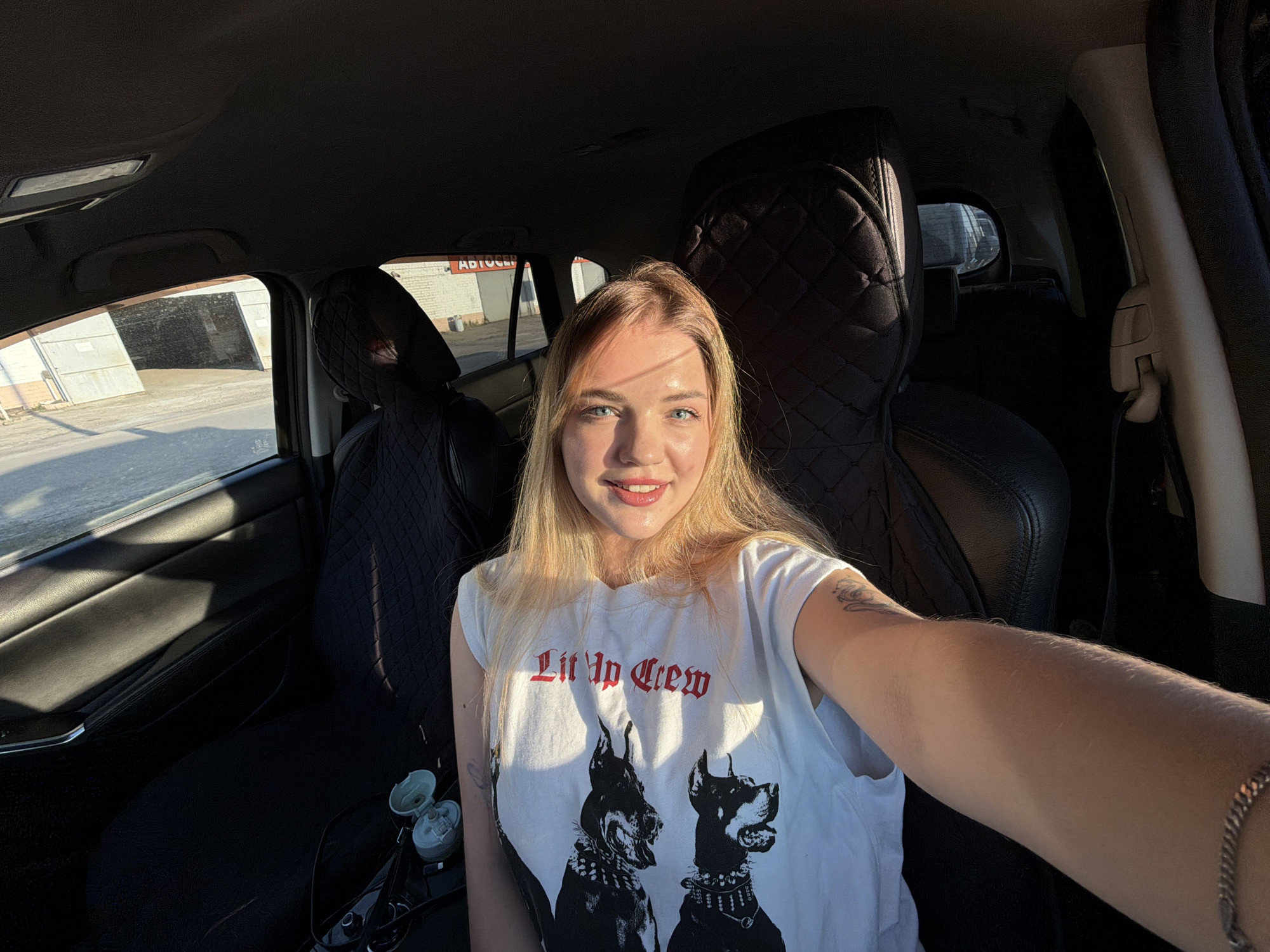 A woman is taking a selfie inside a car. She has blonde hair, blue eyes, and is wearing a white t-shirt with the words "Lit Up Crew" and an image of two dogs. The car's interior is mostly black. The background shows a building with a sign in Russian, partially obscured, but likely advertising "Автосервис" (Auto Service). The location is likely Naberezhnye Chelny, Russia, based on the presence of the Russian language.