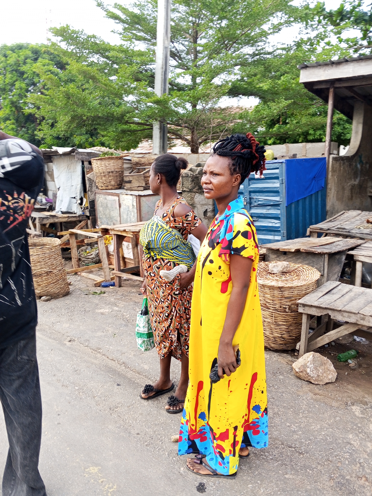 Two women stand on a paved surface in Zango-ndaji, Nigeria. The woman in the foreground wears a long yellow dress with red, blue, and black designs. Her hair is styled in braided buns with red highlights. She holds a dark object in her left hand. The other woman, partially obscured, wears an orange patterned dress and carries a bag. She has a baby wrapped in a striped cloth tied to her body. In the background, there are numerous woven baskets, some on tables. A blue shipping container and other makeshift structures are also visible. A utility pole rises in the background, alongside trees. The area appears to be a marketplace or a commercial zone.