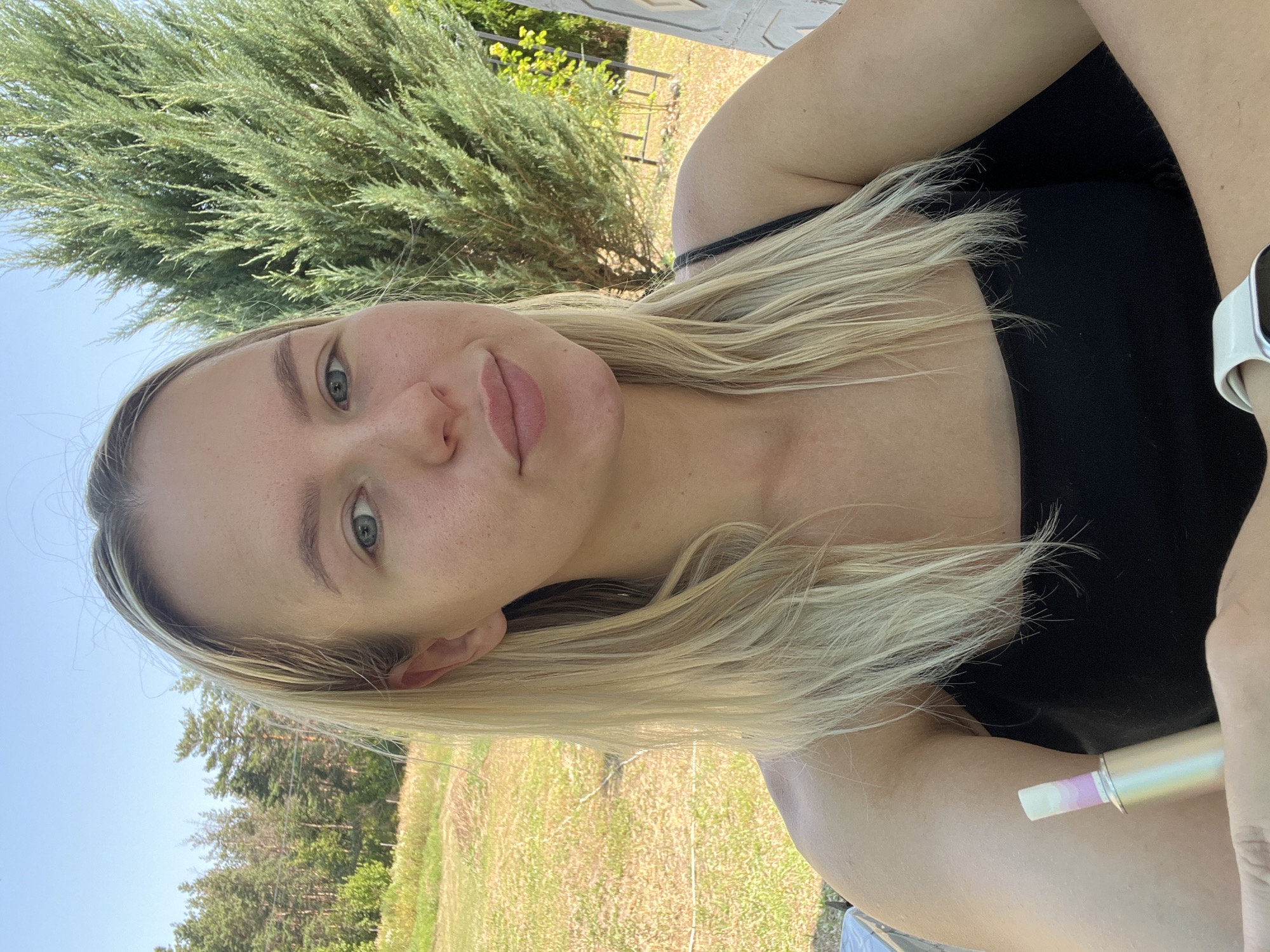 A young Caucasian woman with blonde hair and blue eyes is captured in a close-up selfie outdoors. She is wearing a black tank top. In her left hand, she is holding a device that appears to be a vape. The background suggests a rural or park-like setting with trees, grass, and a clear blue sky. Her arms are crossed, and she is looking directly at the camera. The lighting indicates it is daytime, likely sunny. The image is a selfie, implying the woman is taking the photo herself. The scene is not identifiable as being in a specific city or location.