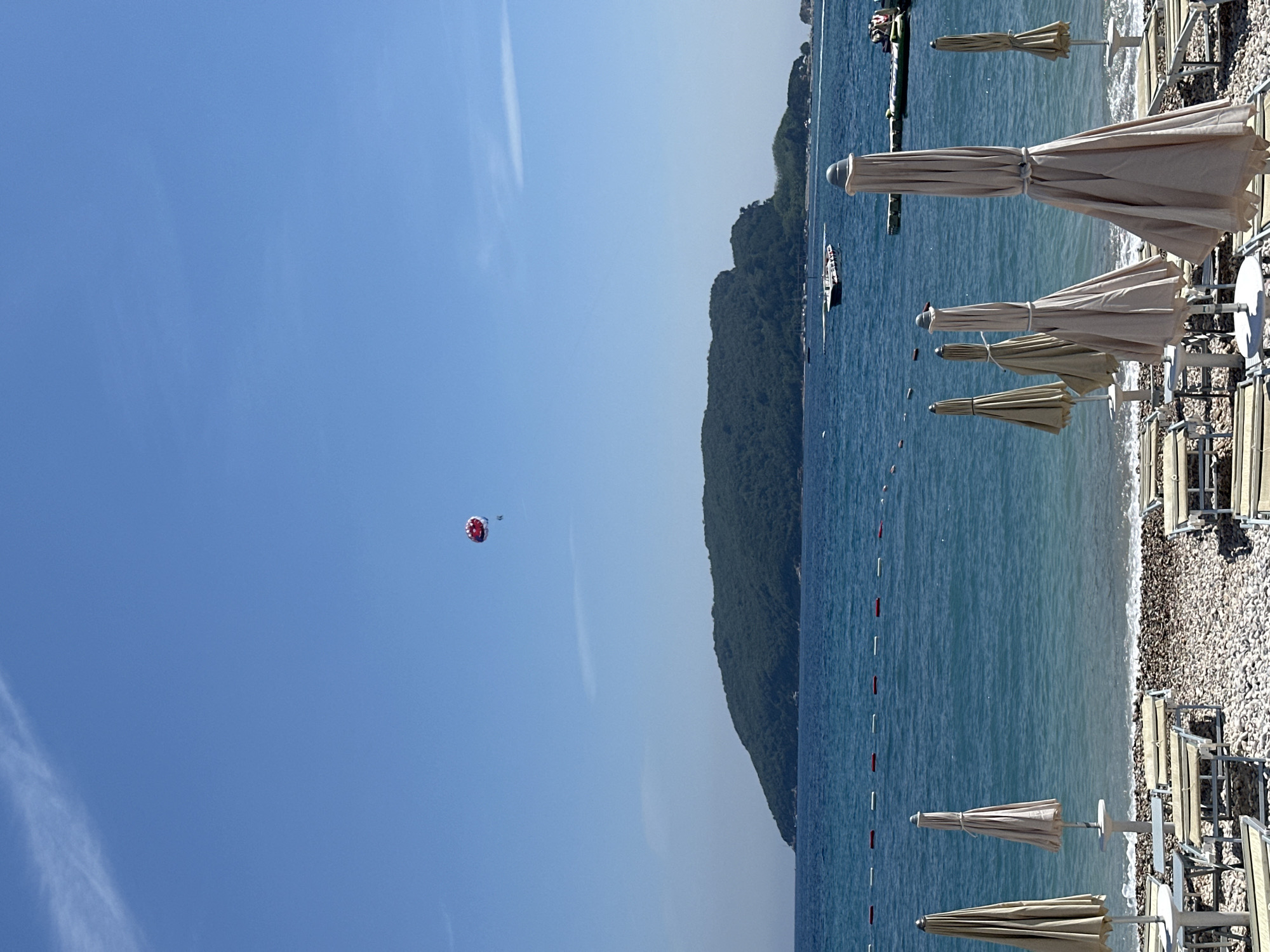 The image, taken on a bright, sunny day in Budva, Montenegro, presents a scenic view of a coastal resort. The foreground on the right features a pebble beach with several rows of neatly folded, light beige beach umbrellas and accompanying lounge chairs. The calm, blue sea stretches across the center and left of the image, with a line of red and white buoys marking a swimming or activity boundary.
In the mid-distance on the left, a large, dark green, tree-covered mountain forms the natural coastline. Several small boats are visible moored in the water near the base of this mountain. The sky above is a vast expanse of clear, bright blue, with a faint wispy cloud visible. High in the upper left portion of the sky, a person is seen parasailing under a striking red and white parachute.
The overall scene suggests excellent weather conditions for beach activities and water sports, indicative of a popular summer tourist destination. The time of day appears to be mid-day due to the bright, direct sunlight. No people are visible on the beach itself. There is no visible text in the image.