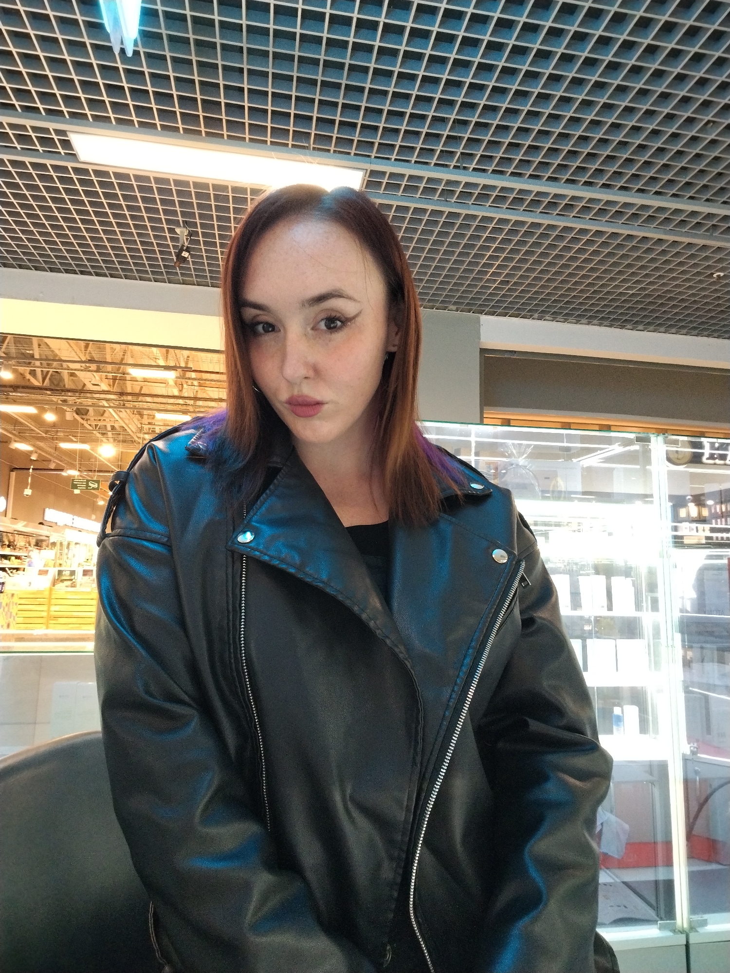 A young woman with reddish-brown hair, featuring subtle purple highlights, poses for a medium close-up photo in an indoor commercial setting in Izhevsk, Russia. She has light skin with freckles, winged eyeliner, and light pink lipstick, looking directly at the camera with a slight pout. She is wearing a black faux leather biker jacket with prominent silver zippers and snaps over a black top.
The background features a grid-patterned dropped ceiling with various rectangular and circular artificial light fixtures. To the left, a bright section of a large store or supermarket is visible with shelves, product displays, and additional overhead lighting, suggesting an extensive retail environment. To the right, clear glass display cases reflect the interior lighting, possibly showcasing merchandise or forming part of a checkout area. A dark, possibly leather, seat is partially visible in the lower left corner. The scene is illuminated by the artificial lighting characteristic of a shopping facility. No discernible text is visible.
