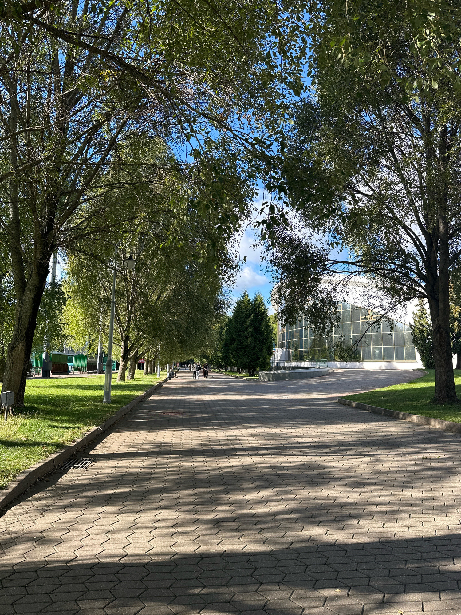 The image depicts a paved walkway in a park, lined with trees on both sides. A modern building with a glass facade is visible in the background. Several people are walking along the path. The scene is well-lit by sunlight, creating dappled shadows on the walkway. The walkway appears to be made of interlocking concrete pavers. The trees are in full foliage, suggesting it is either late spring or summer. The presence of a green utility structure on the left side of the image and the architectural style of the building are consistent with the National Library of Belarus, located in Minsk, Belarus.