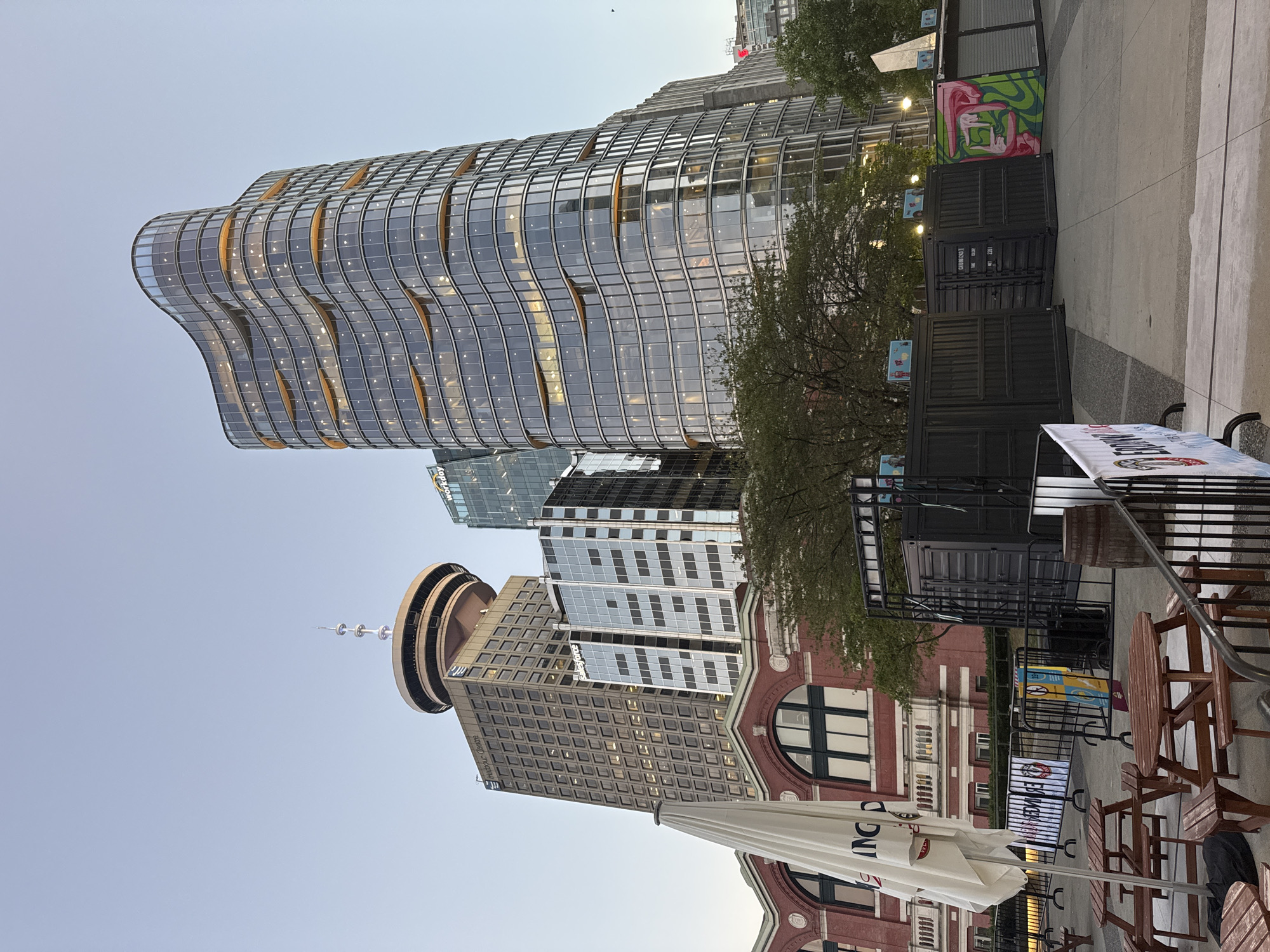 This image captures a street-level view of downtown Vancouver, Canada, focusing on the intersection of modern architecture and public space. Dominating the skyline are several prominent buildings: the Harbour Centre with its distinctive observation deck, a sleek, curvilinear glass skyscraper with illuminated floors, and other contemporary office buildings. In the foreground, a red brick building with arched windows and a white umbrella suggests an outdoor seating area, possibly associated with a restaurant or market. Several black shipping containers, some adorned with graffiti, are arranged in a row, implying a temporary outdoor setup or market stalls. Picnic tables and outdoor seating are visible, indicating a public gathering space. The overall atmosphere is that of an urban environment during twilight, with clear skies and the warm glow of building lights.