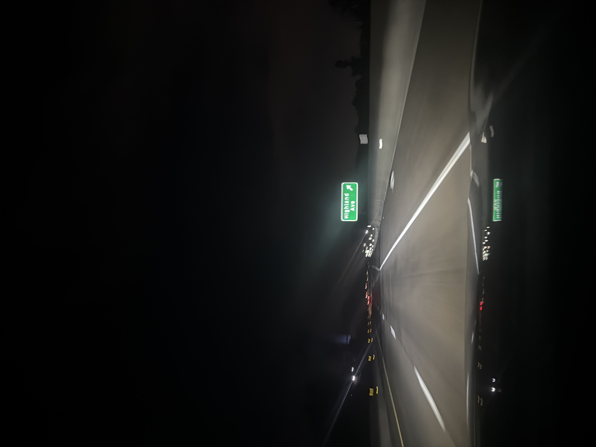 The image depicts a dark, nighttime scene on a highway in Middletown Township, United States of America. A green exit sign indicating "Highland Ave" is visible on the right side of the road. Multiple car headlights and taillights are visible in the distance. White lane markings divide the highway lanes. The left side of the image is mostly dark, with a hint of trees in the far distance.