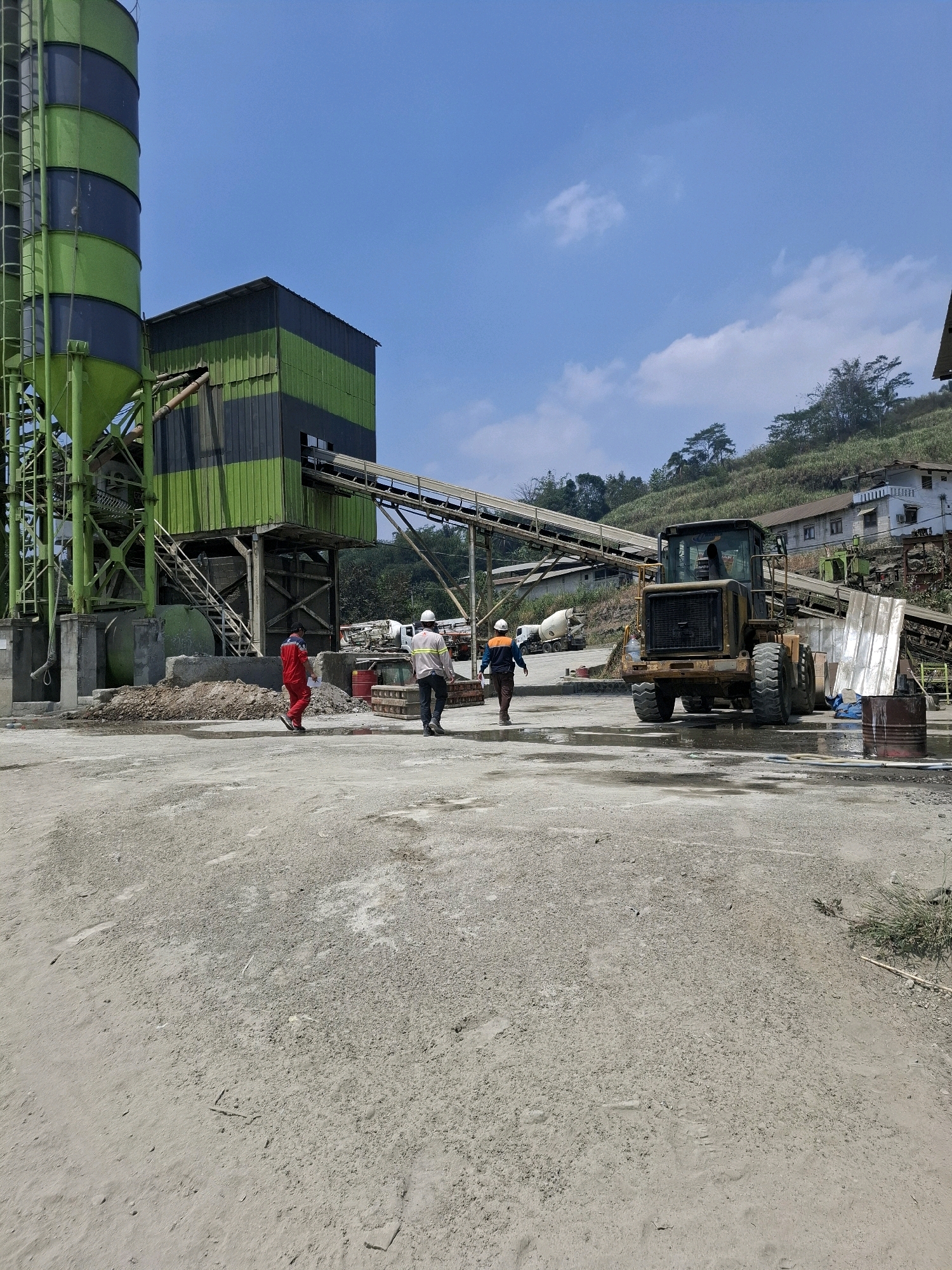 The image depicts a construction site with industrial equipment and workers. In the foreground, a dusty, uneven dirt surface is visible. Two individuals, one in a red uniform and the other in a white hard hat and reflective vest, are walking away from the viewer. To the right, a yellow heavy-duty loader is parked. In the background, a large green and black structure, likely a cement silo, dominates the left side of the frame. A conveyor belt system extends from this structure. Further in the background, a hillside with some buildings and vegetation can be seen under a bright blue sky with scattered white clouds. The scene appears to be in a rural or semi-industrial area, consistent with the location of Cijangkar, Indonesia.