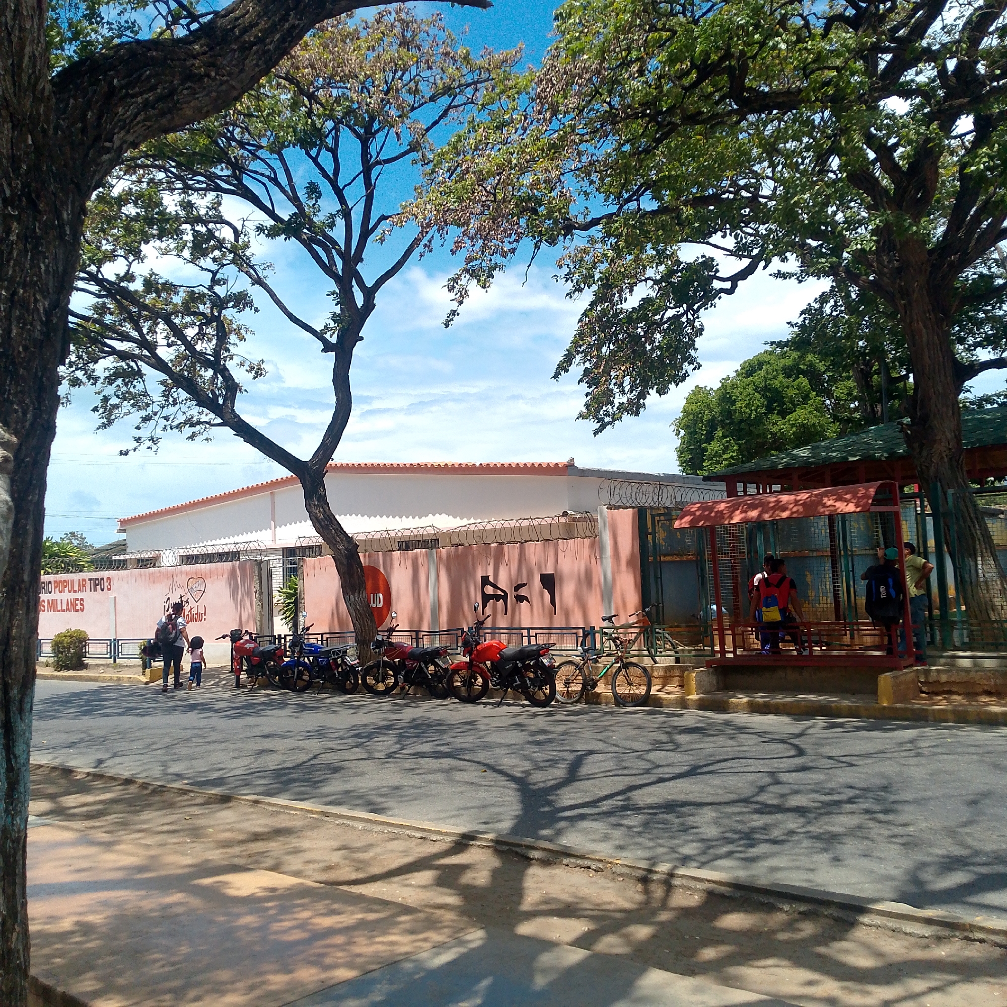 A daytime outdoor shot in Altagracia, Venezuela, shows a street scene with several people and vehicles near a building. A mother and child walk along the sidewalk in front of a light pink building with a white roof. Graffiti decorates the building. Several motorcycles and a bicycle are parked along the curb. A bus stop shelter stands to the right, with several people inside. Trees frame the scene, casting shadows on the street. The sky is blue with scattered clouds.