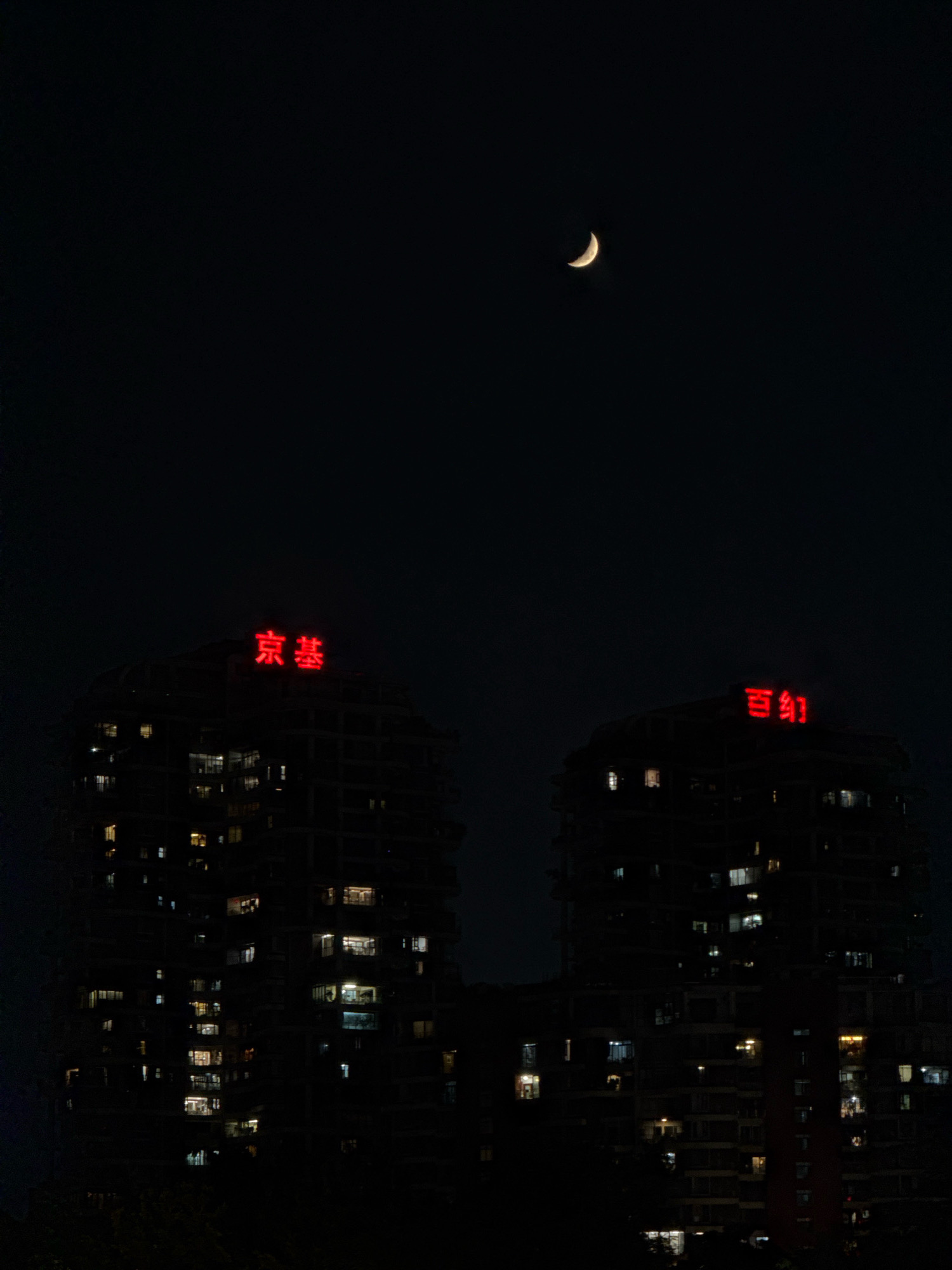 The image depicts a nighttime view of two silhouetted apartment buildings in Nanshan District, China. A crescent moon is visible in the dark sky above. The buildings are illuminated from within, with numerous windows glowing with yellow and white light, suggesting occupancy. Red neon signs are mounted on the rooftops of both buildings. The sign on the left building reads "京基" and the sign on the right building reads "百幻". The foreground is dark and indistinct, with hints of foliage visible at the bottom of the frame, suggesting the viewpoint is from a lower elevation or across a distance. The overall scene is one of a typical urban night environment.