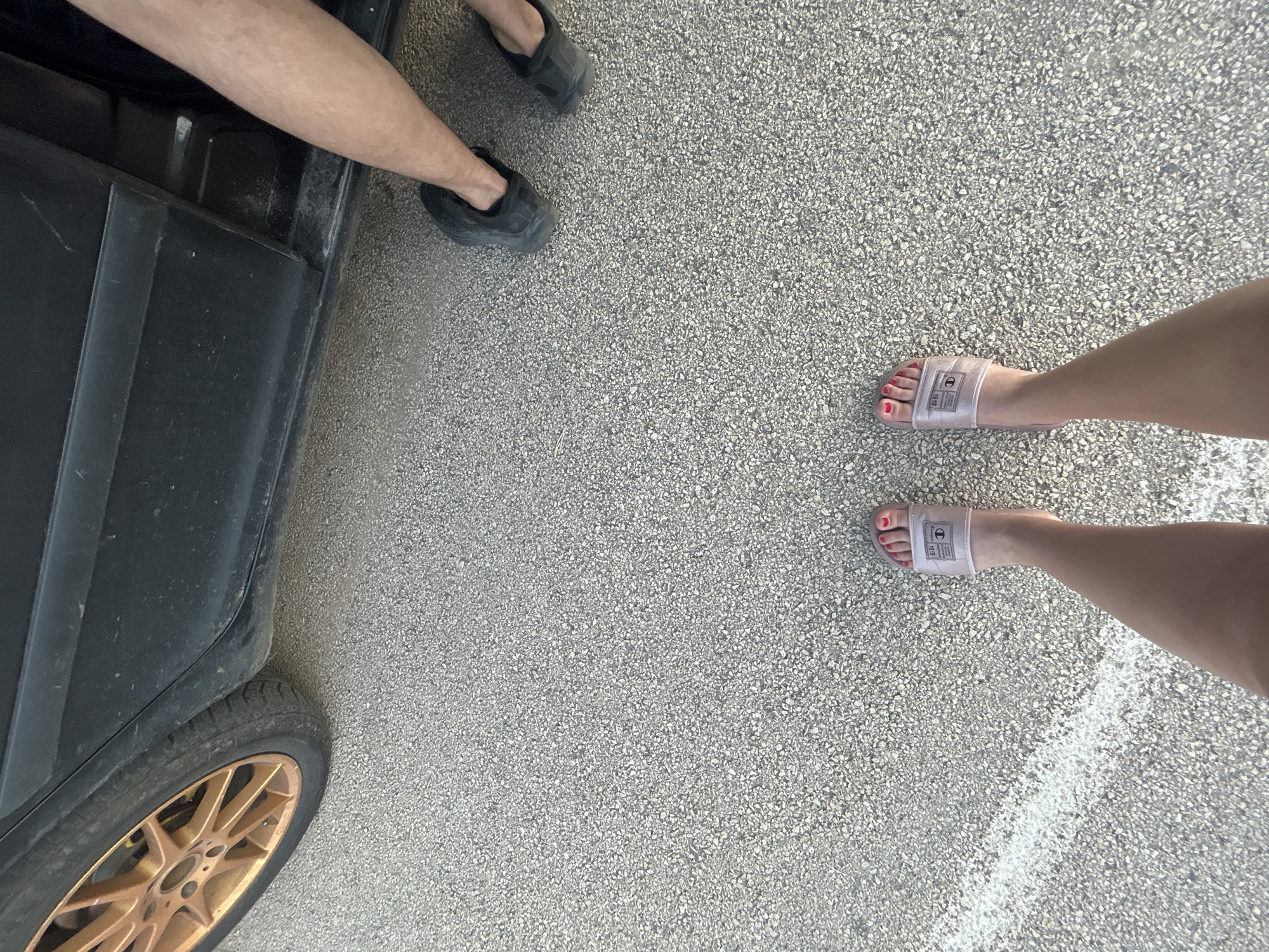The image shows a low-angle shot of two people's legs and feet standing on asphalt. A portion of a black car with a gold rimmed wheel is visible on the left side of the frame. The person on the left has their legs crossed, wearing black slides. The person on the right is wearing clear slides with a logo and red nail polish. A white line is painted on the asphalt in the lower left corner. The scene is an outdoor setting, likely a street or parking lot. No specific landmarks in Knezha, Bulgaria are identifiable.