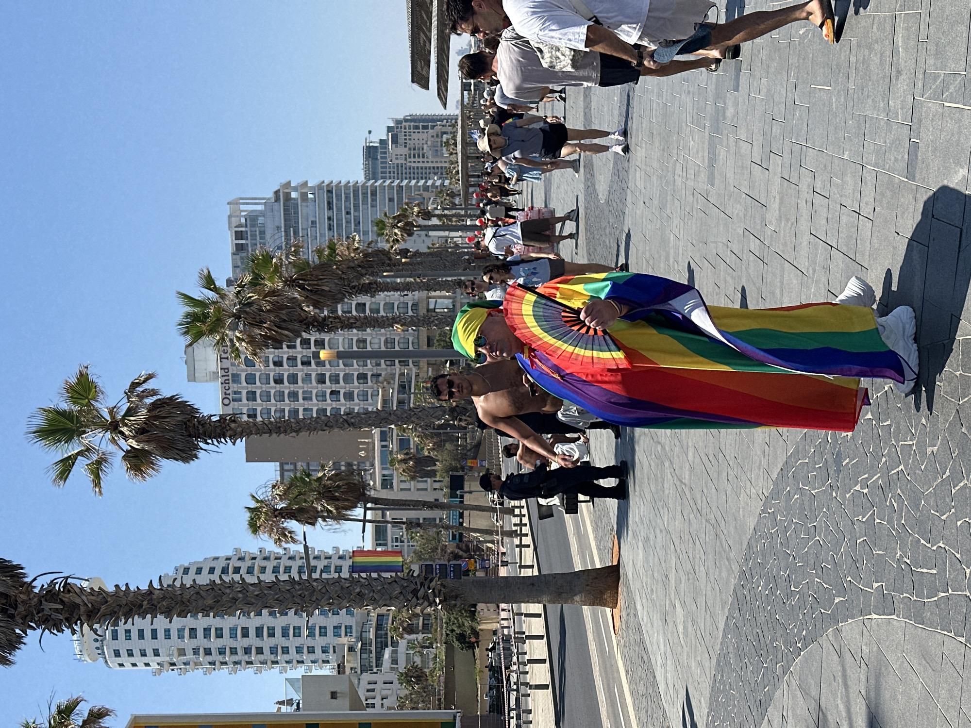 In Tel Aviv, Israel, a person wearing a rainbow-colored robe and a fan stands on a promenade. The person has a yellow bandana around their head and is holding the fan, which is also rainbow-colored, in front of them. Behind this individual, a shirtless man with sunglasses is visible, and further in the background, a crowd of people walks along the promenade. In the distance, tall, modern buildings line the street, with palm trees scattered throughout the scene. A rainbow flag is also visible hanging from a tree on the left side of the image. The weather appears to be sunny with a clear blue sky. The promenade is paved with grey stone tiles, and a street with vehicles is visible to the left of the promenade.