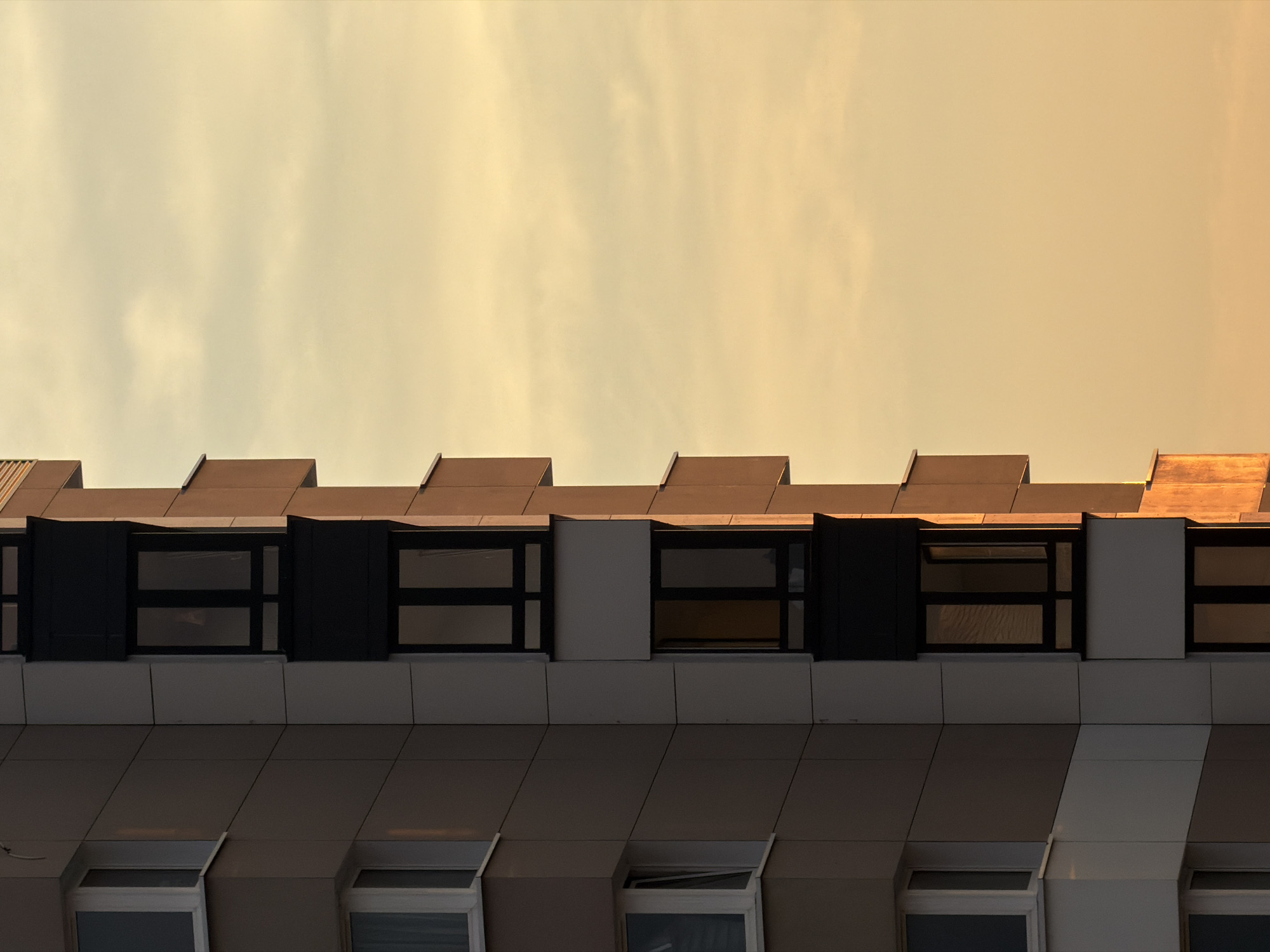 An exterior, medium shot, taken at a slight upward angle, captures the corner of a modern apartment building in Homyel, Belarus, against a softly lit, pale yellow sky filled with hazy clouds. The building's facade, constructed with light brown and white paneling, features a series of dark-framed windows and small balconies stacked vertically. The late afternoon or early evening sun casts a warm, golden light on the building's right side, highlighting the texture of the panels and the edges of the balconies. The left side of the building is in shadow, emphasizing the architectural geometry. The windows, some open, others closed, reflect the muted light of the sky. The overall impression is one of quiet urban structure bathed in gentle, diffused light.