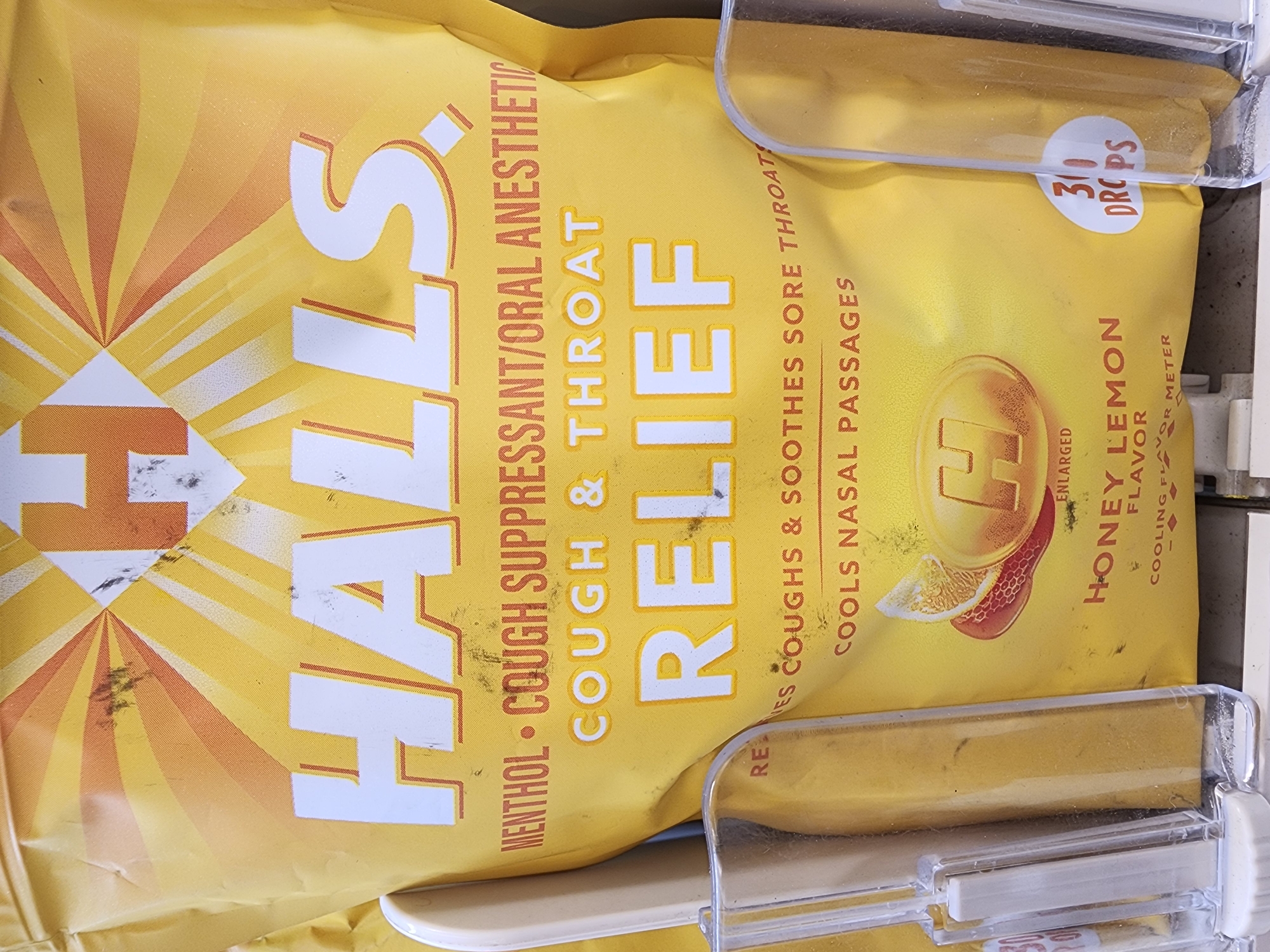 A close-up shot shows a bag of Halls Honey Lemon cough drops. The bag is yellow with orange and yellow sunburst graphics. The brand name "HALLS" is prominently displayed in large white letters with a red outline. Below that, it says "COUGH & THROAT RELIEF" in smaller text. Further down, it states "COOLS NASAL PASSAGES" and features an image of a lemon slice and a cough drop. The text "HONEY LEMON FLAVOR" is also visible. To the right, a circular sticker indicates "300 DROPS". The product is displayed in a clear plastic holder, suggesting it is in a retail environment. There are smudges and dust on the packaging and holder, typical of a store display. The context of Camp Hill, United States of America, is not directly evident from the image itself but would refer to the location where this photograph was taken.