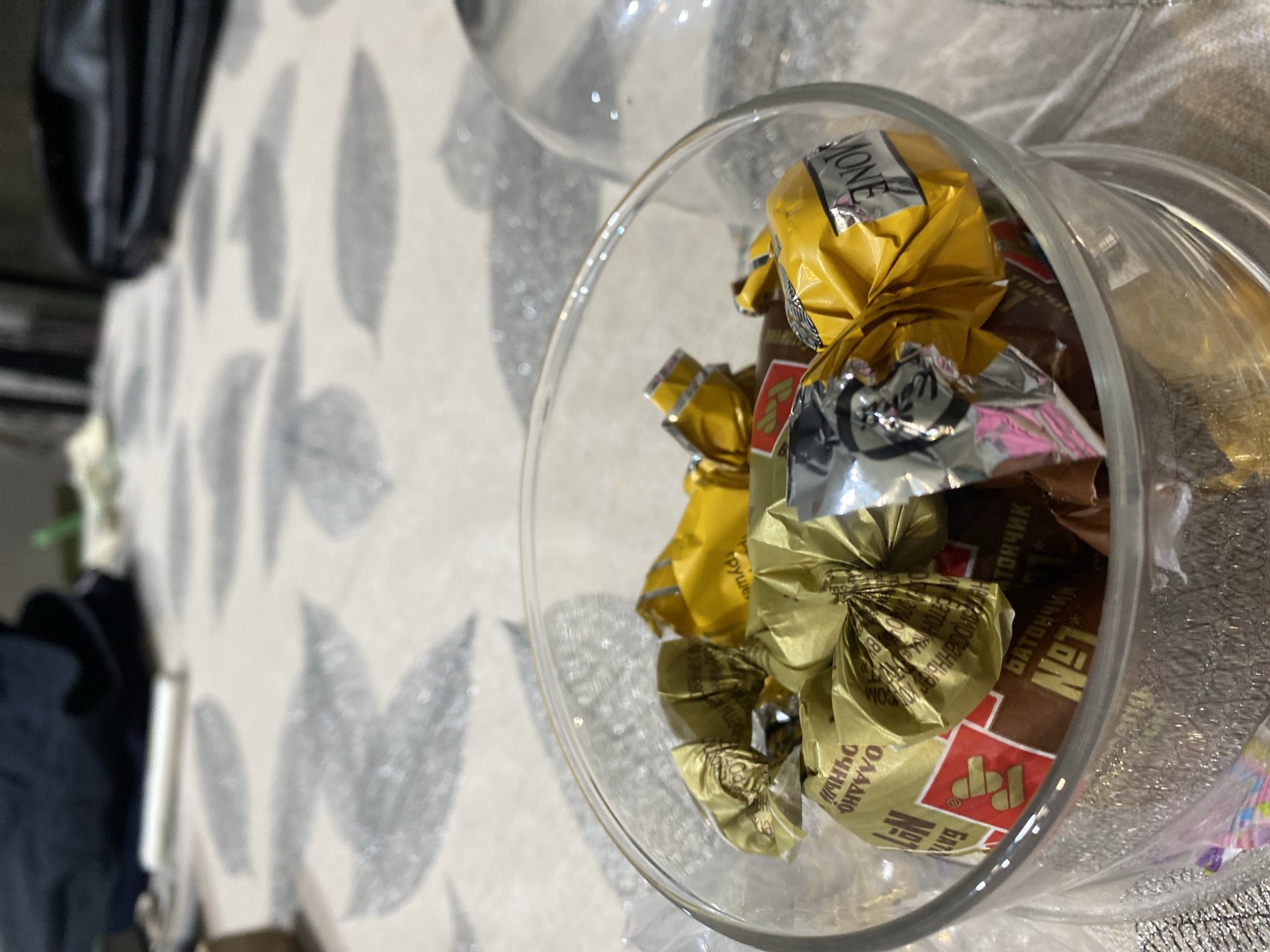 A close-up, high-angle shot shows a clear glass jar filled with wrapped candies on a patterned surface, presumably a tablecloth. The jar is positioned towards the right of the frame. The candies are wrapped in colorful foil, with visible colors including gold, silver, and brown. Some of the wrappers display text in Cyrillic script. Based on the wrapper visible in the lower right corner, the candies appear to be produced by the Perm confectionery factory, potentially located in Chaikovsky, Russia. The background is blurred. The shot is oriented in a way that the top of the jar is at the top of the picture. The image appears to have been taken indoors, likely in a private home.