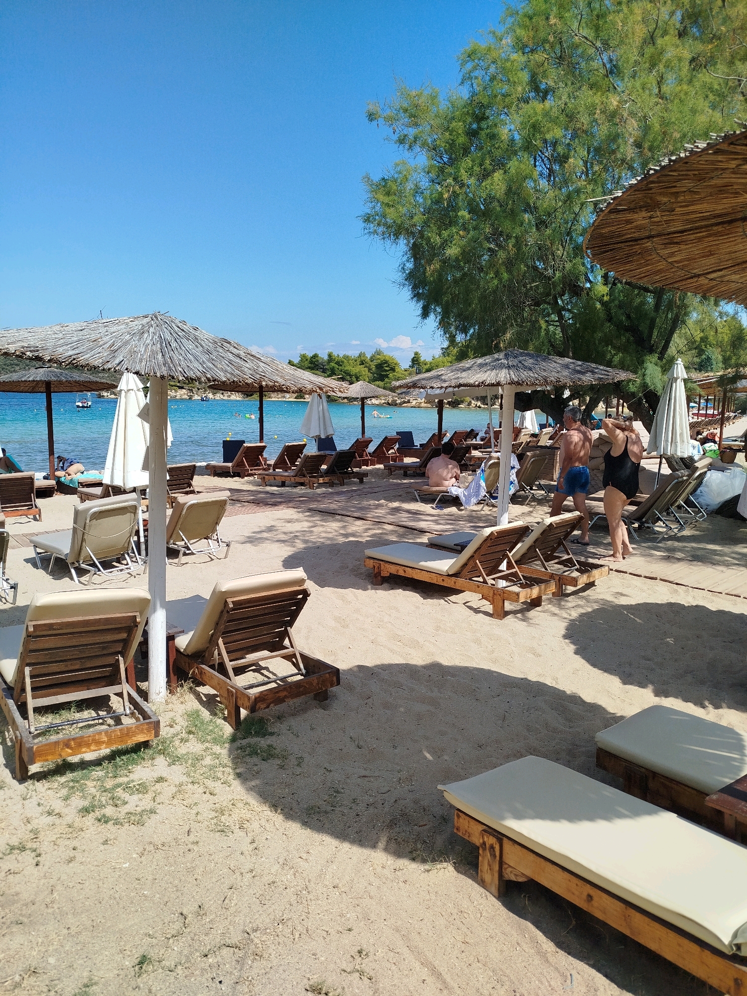 The image depicts a serene beach scene in Sithonia Municipal Unit, Greece. The foreground is dominated by two unoccupied wooden lounge chairs with light-colored cushions, casting distinct shadows on the sandy ground. Beyond these, a variety of other lounge chairs and straw umbrellas are scattered across the beach, some occupied and others vacant. In the middle ground, a man and a woman in swimwear are walking away from the camera. The woman is wearing a black swimsuit, and the man is wearing blue swim trunks. Further in the background, the crystal-clear turquoise waters of the Aegean Sea meet a verdant coastline with lush trees. Several boats are visible on the water, and some people are enjoying the sea. The sky is a vibrant, cloudless blue, indicative of a warm, sunny day.