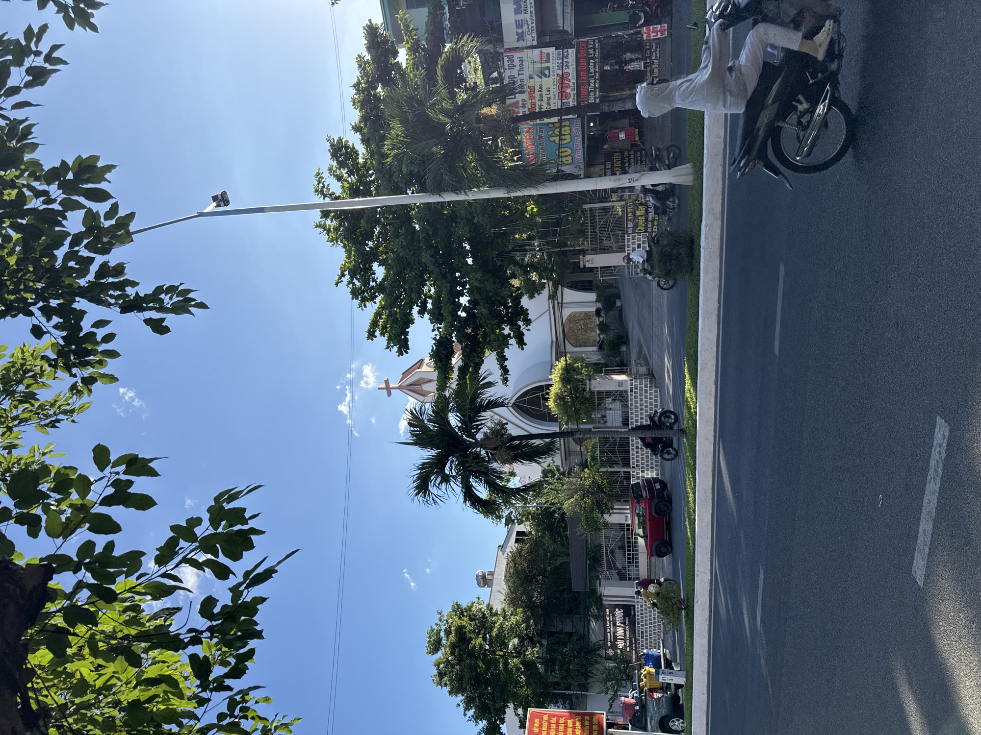 The image shows a street scene in Nha Trang Ward, Vietnam. A two-lane road is visible with trees, buildings, and a church on the left side of the road. There is a person riding a motorcycle on the right-hand side. There are vehicles parked along the road and signs are visible on buildings along the road. The sky is blue with some clouds.