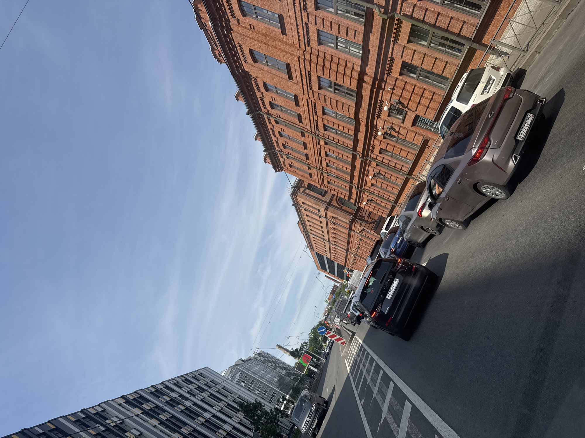 The image depicts a traffic scene on a street in Saint Petersburg, Russia, under a clear blue sky with scattered clouds. On the left side of the street, a modern building with a distinctive facade of dark and light grey panels rises high. To the right of this building, a series of brick buildings with numerous windows line the street, exhibiting a classic architectural style common in Saint Petersburg. The street itself is filled with cars, indicating moderate to heavy traffic. Several vehicles are visible in the foreground and mid-ground, including a dark SUV with the license plate "T415CE 198", a silver sedan with the license plate "B977TP 147", and other cars further down the road. Traffic lights and road signs are also present, suggesting the area is a busy intersection. Power lines are visible strung across the sky. The perspective suggests the photo was taken from within one of the vehicles, looking forward and slightly to the right.