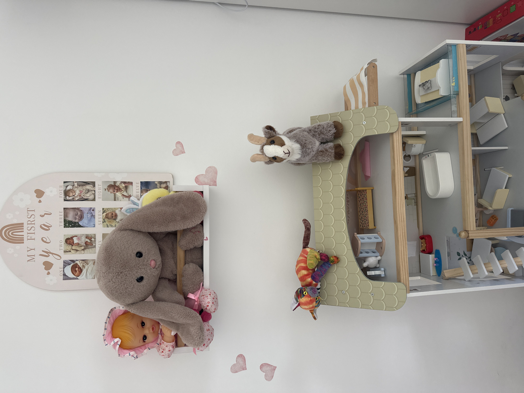 A children's room in Petah Tikva, Israel, displays a dollhouse and plush toys against a white wall adorned with decorative pink hearts. A large, gray plush bunny is perched on a white shelf, partially obscuring a vintage-style doll with orange hair and a pink bonnet. To the left of the bunny, a pink floral fabric is visible. Above the shelf, a wooden plaque displays a rainbow and the text "MY F<bos> LSRST year" with framed baby photos.
On top of the dollhouse, a small, plush goat with brown and white fur stands facing forward. A colorful, striped plush cat with a segmented body is draped over the dollhouse roof. The dollhouse itself is a multi-story structure with visible rooms and furniture, including beds, a bathtub, and other miniature items. A white wooden ladder leads to the upper levels of the dollhouse. To the right of the dollhouse, a portion of a colorful toy or game with red and yellow elements is visible. The lighting suggests an indoor environment, likely during the daytime.