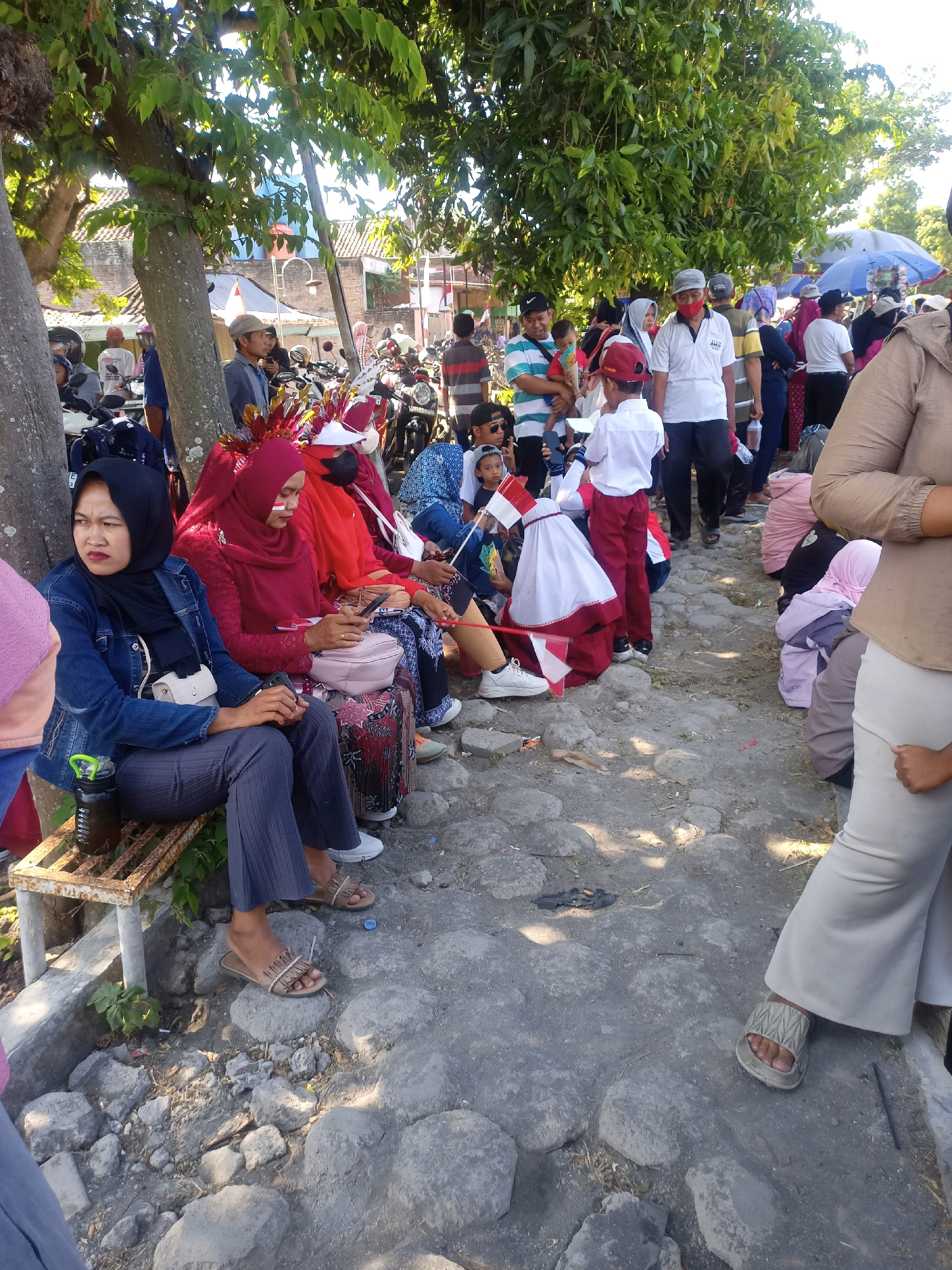 A group of people, some dressed in traditional Indonesian attire, are gathered outdoors in what appears to be a street or public area in Sumberlawang, Indonesia. The scene is illuminated by bright sunlight, casting shadows on the rocky ground. Several motorcycles are parked in the background, suggesting this might be a local event or gathering. Some individuals are seated on a bench, while others are standing or walking. A few children are present, one of whom is dressed in a uniform with a red and white flag. The overall atmosphere seems relaxed and communal, with many people wearing masks, likely due to health precautions. The environment includes trees providing shade, buildings in the distance, and various onlookers.