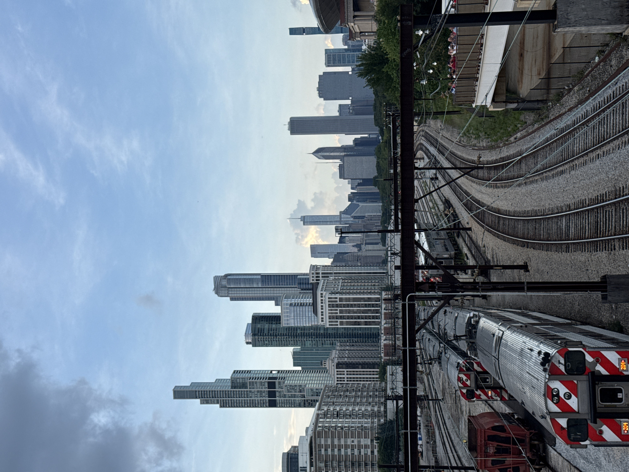 This is a high-angle, medium shot photograph of a cityscape with trains in the foreground, taken from a bridge or elevated walkway. The scene is set in Chicago, Illinois, United States of America.
In the foreground, multiple train tracks curve into the distance, with several commuter trains parked or moving along them. The trains are silver with red and white stripes on the front. To the left, a red freight car is visible. An elevated walkway with a railing stretches across the mid-ground to the right, where a group of people, some wearing red clothing, are standing and looking out.
In the background, the Chicago skyline is visible with numerous skyscrapers of varying heights and architectural styles. The sky is partly cloudy with dramatic, dark clouds on the left and lighter, brighter clouds on the right. Lush green trees are visible between the train tracks and the skyline, and also to the right of the tracks, near the walkway. The overall lighting suggests it is late afternoon or early evening, with a muted quality due to the overcast sky.