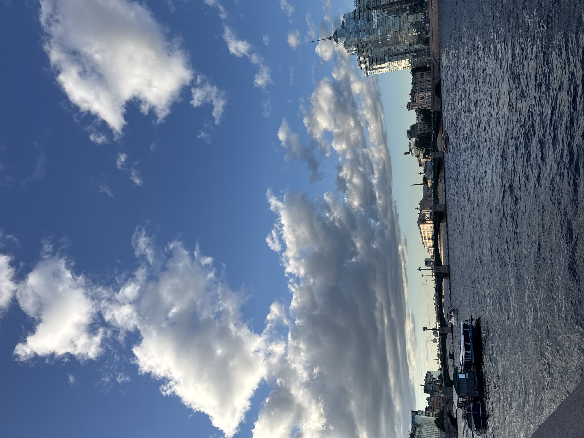 This is a full-frame, eye-level shot of the Neva River in Saint Petersburg, Russia. The sky is a vibrant blue, dotted with numerous white and gray cumulus clouds, some of which are quite large and dramatic. The river takes up the bottom half of the frame, its dark blue waters rippling.
On the left bank of the river, several boats are docked or moving. One larger boat, a tour boat with a cabin and an open deck area, is prominent. It appears to be carrying passengers. Another smaller dark blue boat is tethered nearby. Further down the river, a modern, multi-story building with a glass facade and a spire is visible on the right bank, reflecting the sky.
In the middle ground, a long bridge with multiple arches spans the river. Cars can be seen crossing the bridge. Beyond the bridge, a cityscape with various buildings, including some older, ornate structures and more modern ones, stretches along the riverbank. The lighting suggests it is late afternoon or early evening, with the sun casting a warm glow on some of the buildings and clouds. The overall scene depicts a busy urban waterway with elements of both historic and modern architecture.