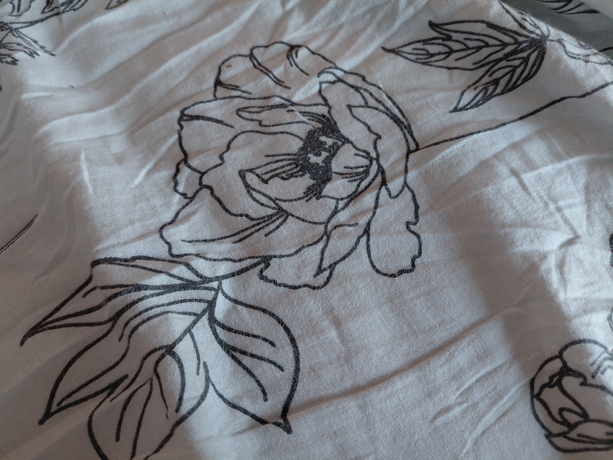The image displays a close-up view of a white fabric with a black floral print. The fabric is creased and textured, with visible wrinkles and folds. The floral pattern features stylized flowers and leaves outlined in thin black lines. One prominent flower is in the center of the frame, with delicate petals and a detailed stamen. Several leaves are also visible, some with distinct vein patterns. The overall impression is one of a soft, draped textile with an artistic, minimalist design. There are no people, actions, or environmental elements present in the image, as it is a close-up of a material. The location in Malinovka, Russia is not visually represented in this image.