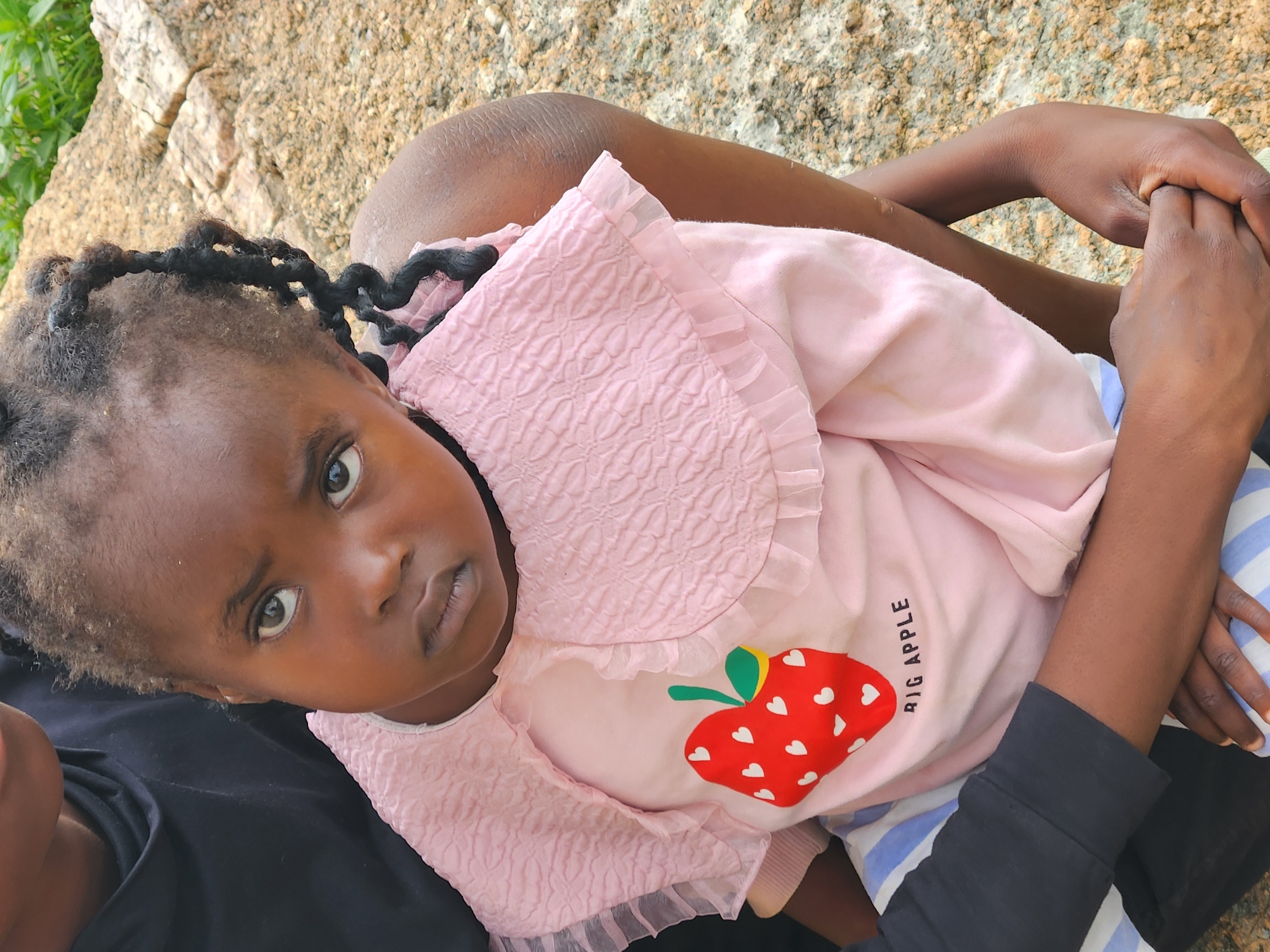 A close-up, eye-level shot features a young Black girl with braided hair, wearing a pink ruffled shirt with a strawberry graphic and the words "BIG APPLE" printed below it, and striped pants. She is being held by an adult, whose dark clothing is partially visible. They are seated outdoors on a rocky surface, with green foliage in the background. The overall scene suggests a casual, possibly familial moment in an outdoor setting. The photographer has likely captured this image within the city of Jos, Nigeria, a location known for its rocky terrain and outdoor spaces.