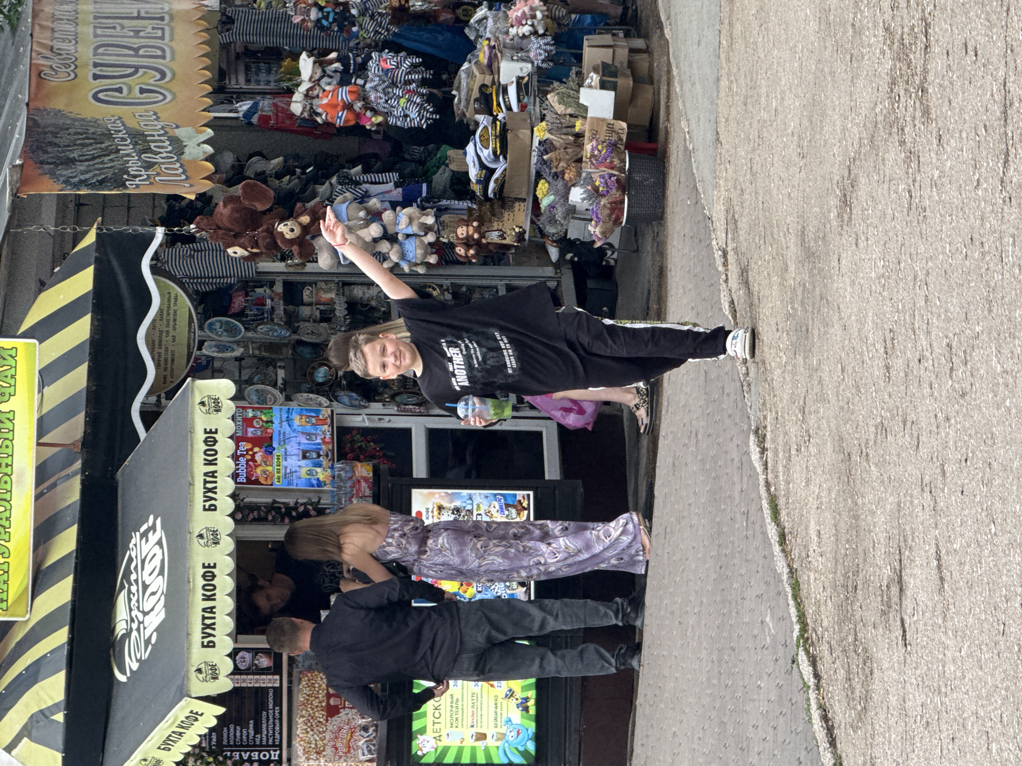 The image depicts a street scene in Inkerman, Ukraine, with a focus on a marketplace or kiosk area. In the foreground, a young boy wearing a black t-shirt and black track pants with white stripes walks towards the viewer, holding a plastic cup with a green beverage and an outstretched arm. Behind him, to the left, a couple is engaged with a vendor at a coffee kiosk labeled "БУХТА КОФЕ" (Bukhta Coffee). The woman is wearing a long, patterned dress, and the man is in a black shirt and jeans. The kiosk displays a menu and various drinks. The background reveals a stall selling an assortment of items, including stuffed animals, hats, and dried flowers, with a banner advertising "Крымская Лаванда" (Crimean Lavender). The environment suggests a bustling outdoor market.