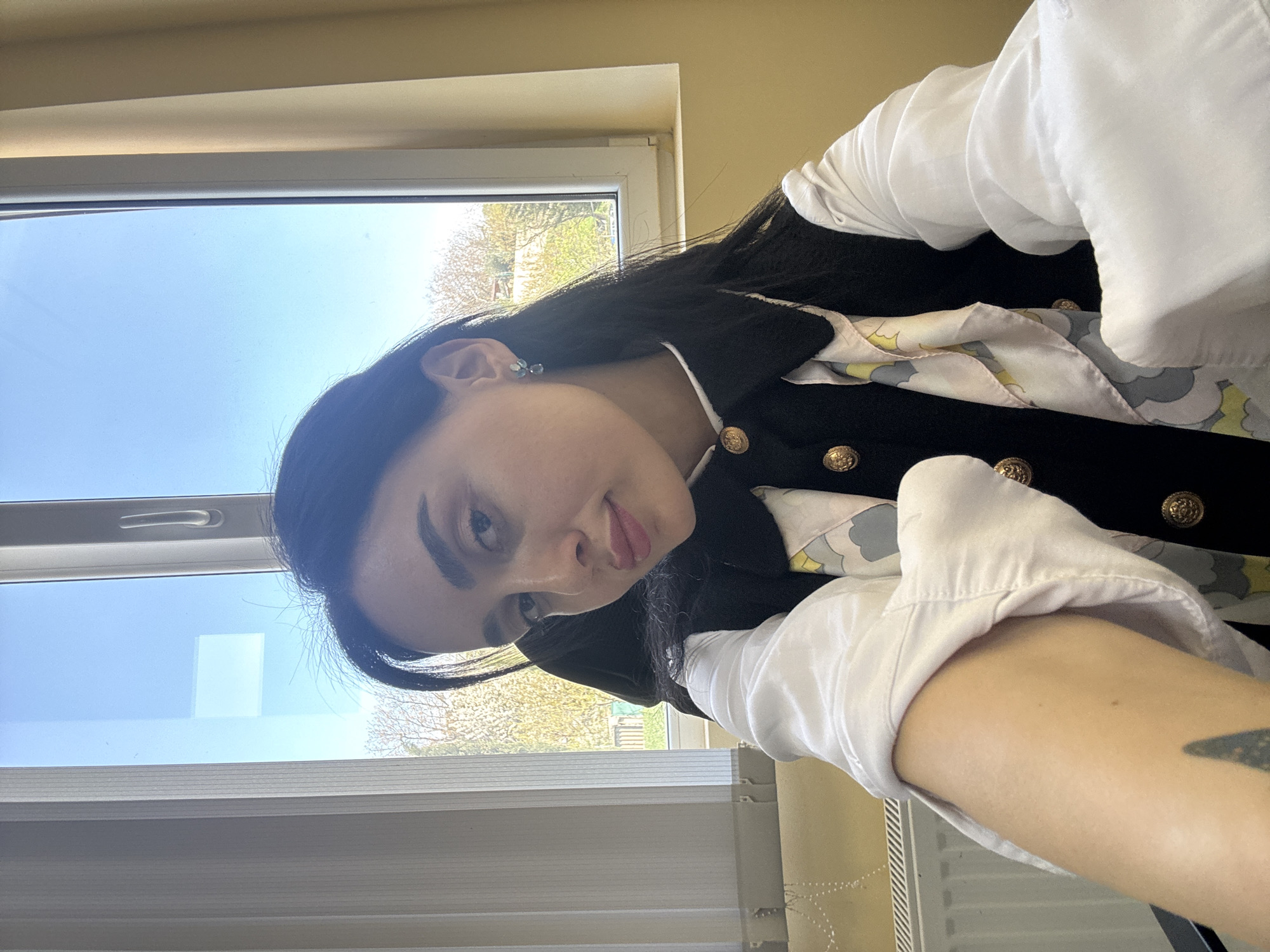 The image features a young woman with long dark hair, looking towards the camera with a neutral to slightly pleasant expression. She is wearing a white long-sleeved shirt layered under a dark vest or jacket with prominent gold-colored buttons. A patterned scarf in shades of light yellow, grey, and white with abstract or cloud-like designs is visible around her neck. A small, light-colored gemstone earring is visible in her left ear. Part of her arm, showing light skin and a partial tattoo, is also visible in the foreground.
The setting is indoors, positioned next to a large window. Through the window, a clear blue sky and sunlit green trees are visible, indicating a bright, sunny daytime, likely during spring or summer. The white frame of the window and horizontal blinds are prominent. A white radiator is partially visible in the bottom right corner of the frame. A white rectangular reflection is present on the windowpane. The overall context for the image is Tbilisi, Georgia. There is no visible text in the image.