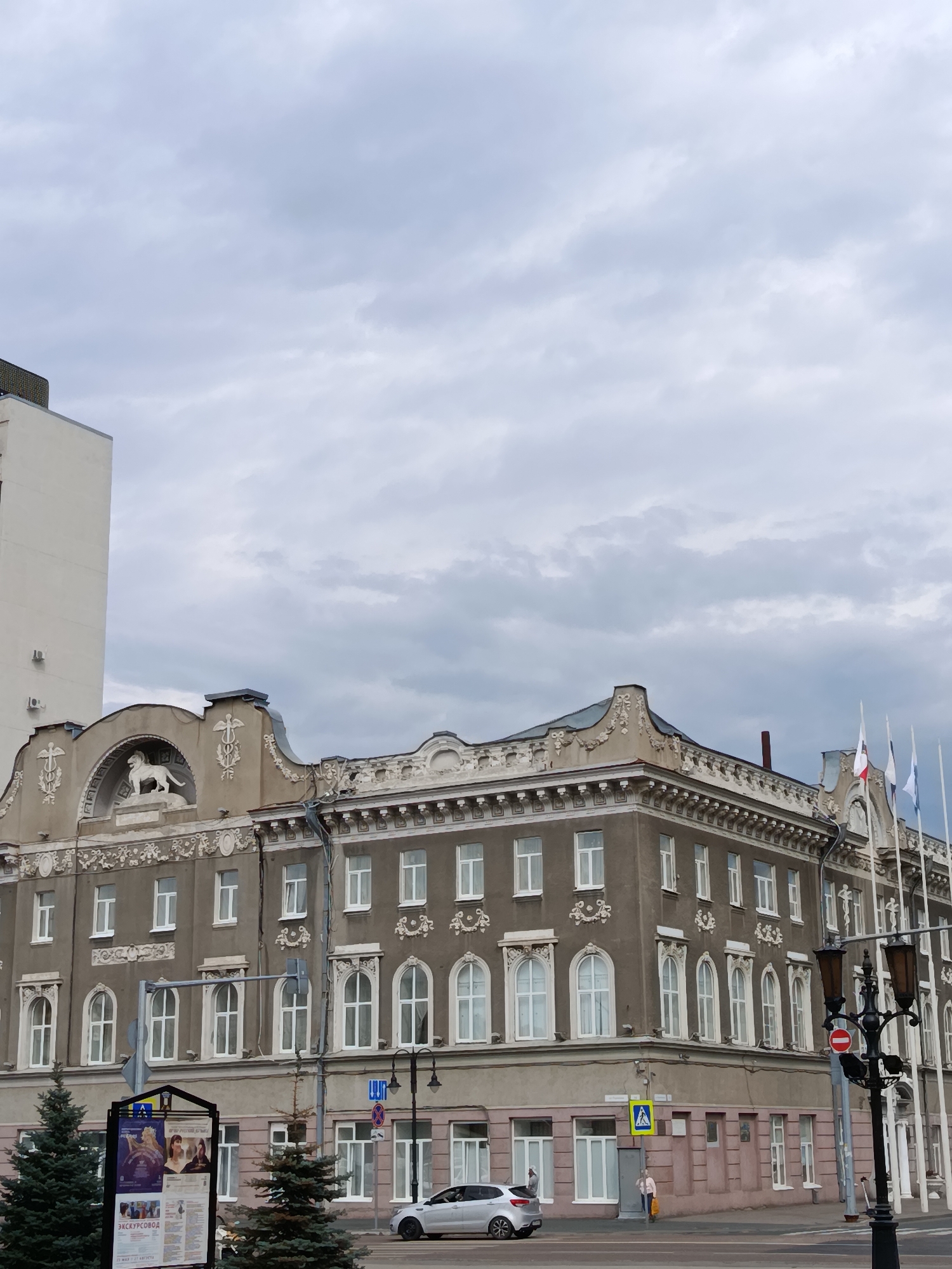 The image depicts a view of a grand, old building in Saratov, Russia, under a cloudy sky. The building, with its ornate facade and mansard roof, features decorative elements such as a sculpted lion, floral motifs, and arched windows. Flags fly from poles on the right side of the building. In the foreground, a street scene unfolds with a silver car, a lamppost, and a billboard displaying posters, one with Cyrillic text that appears to advertise an excursion. A couple of people are visible near the entrance of the building and on the sidewalk. The weather appears overcast, suggesting it might be daytime with diffused light. The architectural style and the presence of Cyrillic text strongly indicate a location within Russia. The overall impression is of a historical urban setting.