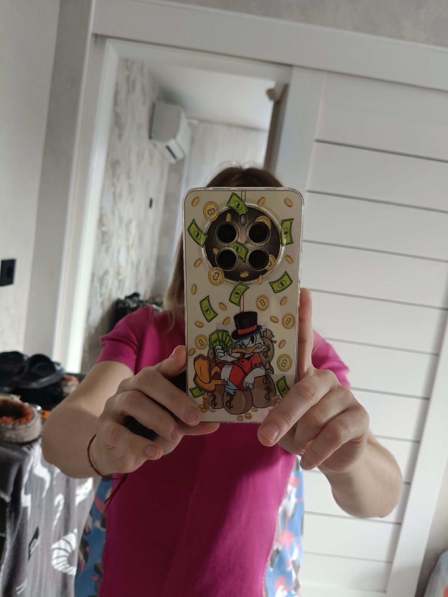 The image shows a person holding a smartphone with a cartoon character and money printed on the back of the phone case. The person is wearing a pink t-shirt. The background appears to be an indoor setting, possibly a room with white walls and a door. There is no discernible connection to the city of Oryol-Izumrud, Russia.