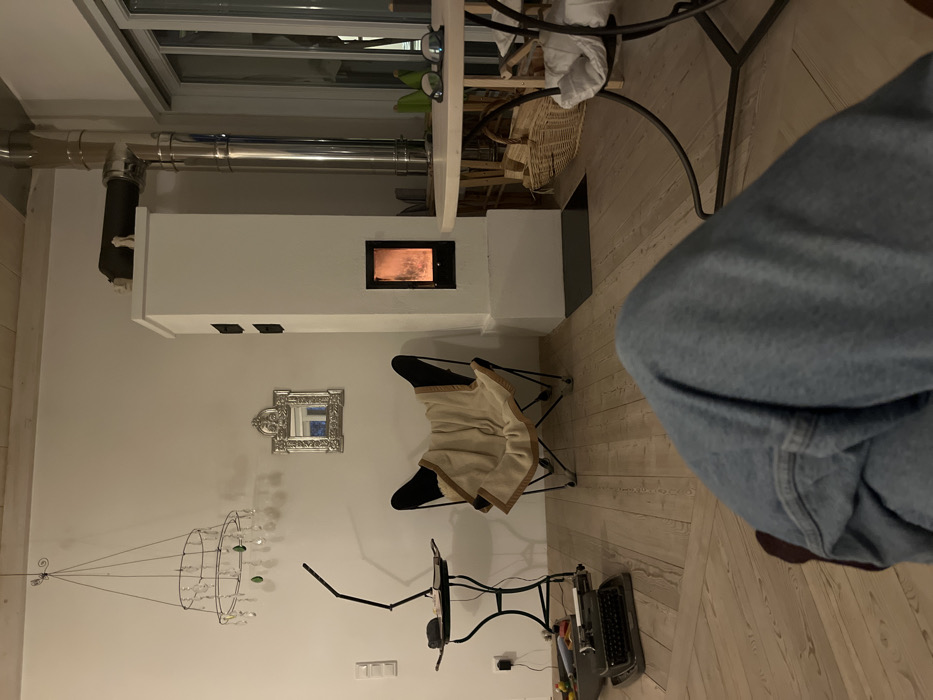 An interior scene within a residential living area in Hohenbrunn, Germany, is depicted from a low, first-person perspective. The lower portion of a person's legs, clad in light blue denim jeans, occupies the immediate foreground, with the left leg bent and partially obscuring dark footwear.
The flooring consists of light-colored wooden planks. To the left, a dark-colored vintage typewriter rests on the floor, alongside several small, colorful objects. Adjacent to this, a black metal side table with an adjustable black desk lamp holds a dark object on its surface. A white electrical outlet is visible on the wall behind.
Centrally positioned is a black butterfly-style chair draped with a light beige blanket featuring a brown trim. Above the chair, an ornate silver-framed mirror reflects a blue-toned image, and a wire-frame chandelier-like decoration with clear and green crystal droplets hangs from the light-colored wooden ceiling.
A prominent feature is a tall, white masonry stove (Kachelofen) located in the midground, with a black-framed firebox opening revealing active orange flames. Two black rectangular access panels are visible higher on its side. A polished stainless steel chimney pipe system extends from the top of the stove, bending towards the right and ascending out of frame.
To the right of the stove, a round wooden table with a black metal base displays a pair of dark-framed glasses and green decorative objects on its surface. A white cloth is draped over part of the table's base. Behind and beneath this table, several wicker baskets are arranged, and a large window or glass door reflects interior light. The background wall is uniformly light-colored. The overall lighting is dim and warm, indicating an indoor evening setting.