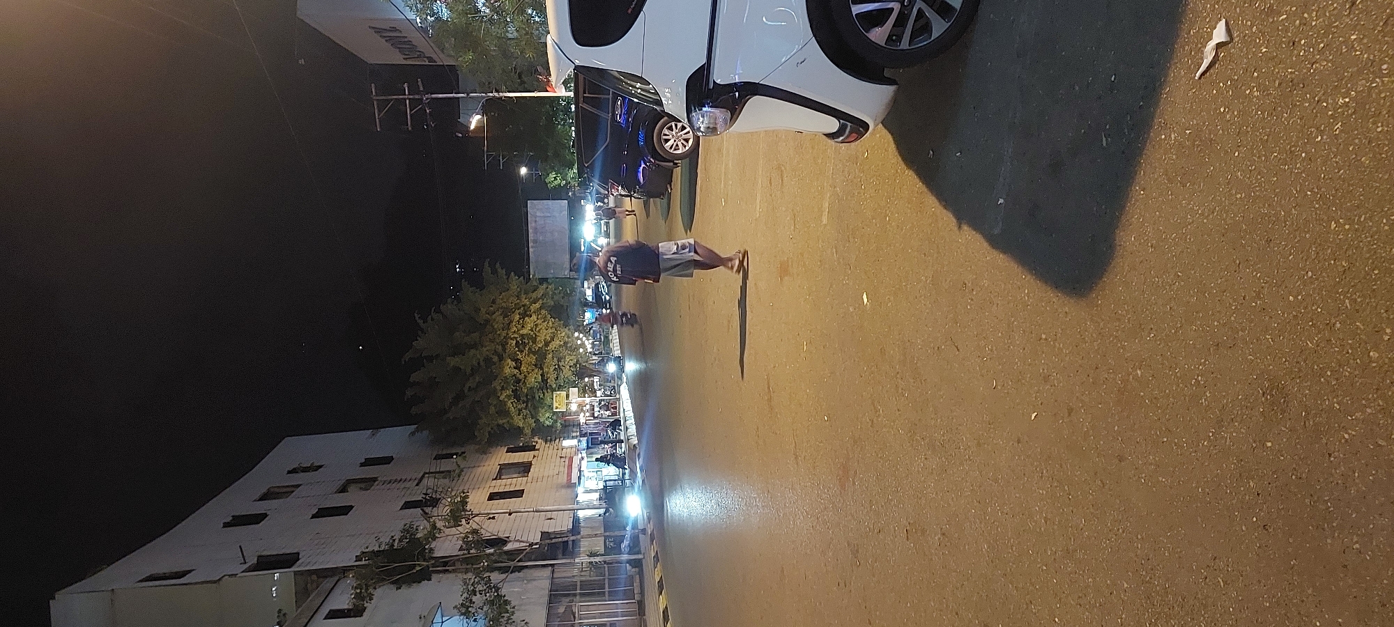 The image depicts a street scene at night in Jayapura, Indonesia, likely in an urban commercial area. A man wearing a dark t-shirt with text and light-colored shorts is walking away from the camera in the middle of the street. He is casting a shadow on the asphalt road. On the right side of the image, the front of a white vehicle is visible, parked or stopped. Further down the street, several other vehicles, including a dark SUV and motorcycles, are present. The street is lined with buildings on the left, some appearing to be commercial establishments with illuminated storefronts. Trees and streetlights are also visible, illuminating the scene. The sky is dark, indicating nighttime, and there are some distant lights suggesting other parts of the city or stars. The overall impression is of a lively but dimly lit street with active traffic and pedestrian presence.