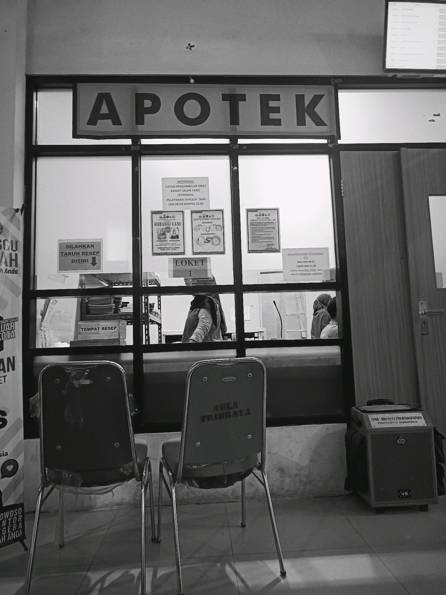The image shows the exterior of an "APOTEK" (pharmacy) in Bondowoso, Indonesia. The pharmacy has a large glass window displaying various signs and posters. Inside, two individuals, presumably staff, are visible behind the counter. One is attending to something on the counter, while the other, wearing a headscarf and long sleeves, is looking down. Two empty chairs, covered in plastic, are positioned in front of the pharmacy entrance, suggesting a waiting area. To the right of the entrance, a portable speaker labeled "V8 BHAYANGKARA PORTABLE MANPACK" is placed on the floor. The overall setting appears to be an indoor space with a tiled floor and bright lighting. The image is captured in black and white, lending it a vintage or documentary feel.