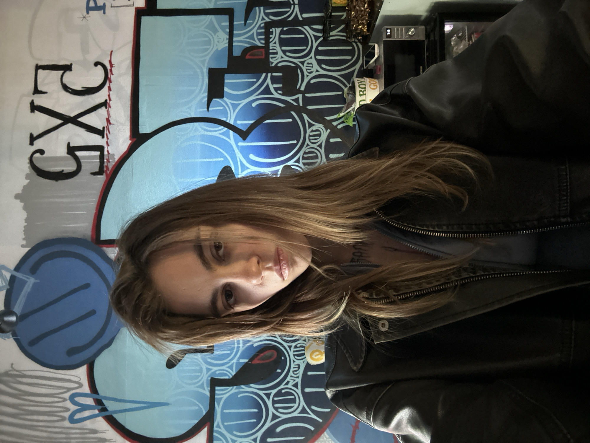 A young woman with long, brown hair and brown eyes, wearing a dark leather jacket over a blue shirt, poses for a selfie in front of a graffiti-covered wall. The graffiti features large blue and grey shapes, along with black and red lettering, including the letters "GXG". A microwave and a plant are visible on a shelf to the right of the woman. The scene appears to be indoors, possibly an urban dwelling or studio space, with no indication of a specific location within Moscow, Russia.