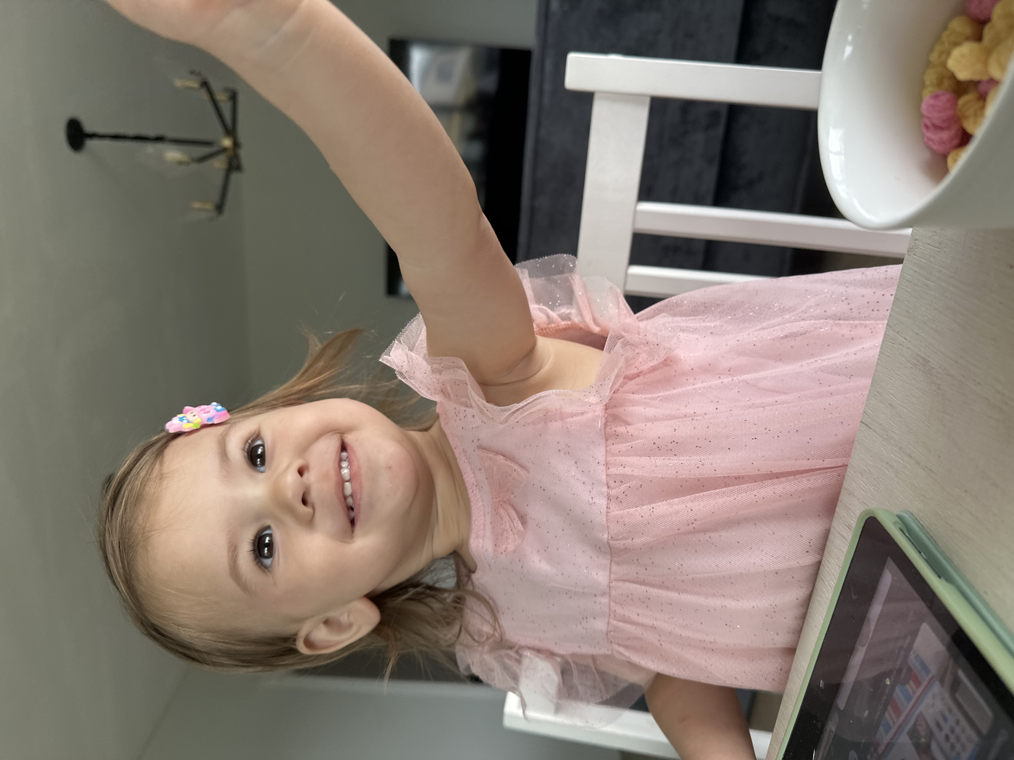 A young girl, likely a toddler, is seated at a table indoors. She is wearing a pink dress with a glittery overlay and a colorful clip in her hair. She is smiling and has one arm raised. A bowl of cereal and a tablet are on the table in front of her. A white chair and a chandelier are visible in the background. It is not possible to determine if the location is in the city of Kharnavy, Russia from the given image.