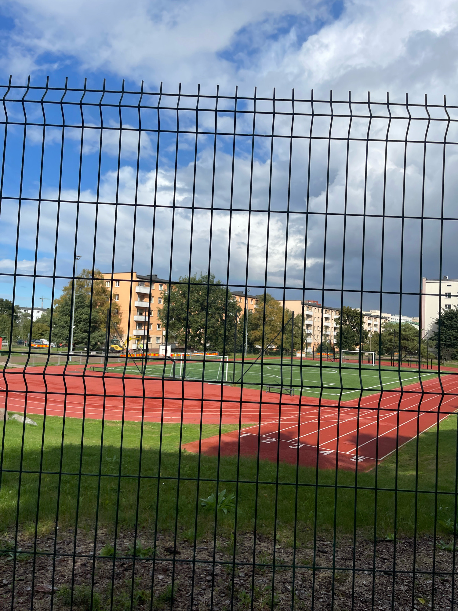 The image shows a fenced-in outdoor athletic facility, likely a stadium, in Tallinn, Estonia. The foreground is dominated by a black chain-link fence. Beyond the fence, a red running track with white lane markers is visible, with lane numbers 2, 3, 4, and 5 clearly marked. Adjacent to the track is a green field, possibly for soccer or other field sports, with a white goalpost. In the background, several multi-story apartment buildings are visible, some with light orange facades and others with white. Trees with green and yellow foliage are interspersed between the buildings and the athletic field, suggesting it is autumn. The sky is partly cloudy with dramatic dark storm clouds and patches of blue sky, and a faint rainbow is visible on the left side of the image. The scene appears to be a public sports area within a residential district of the city.
