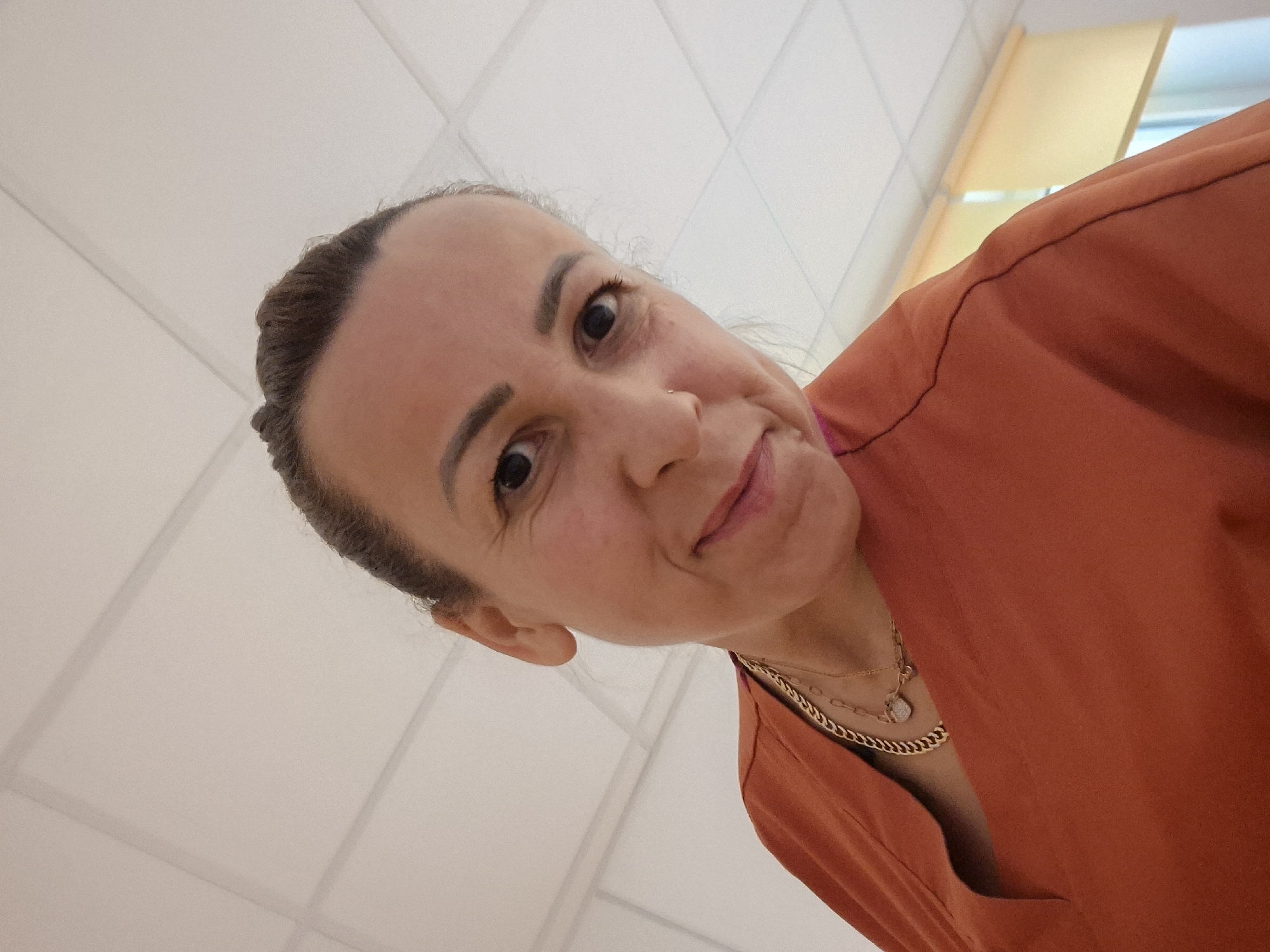 An adult woman with dark hair pulled back and a fair to medium skin tone is depicted in a low-angle, head-and-shoulders shot, looking directly into the camera with a gentle smile. She has defined eyebrows, dark eyes, and a small stud nose ring on her left nostril. She is wearing a rust-orange or terracotta-colored V-neck top. Two gold-toned necklaces are visible around her neck: a thicker curb chain and a thinner chain featuring a small, irregularly shaped pendant. The background consists of a white suspended ceiling with a grid pattern of acoustic tiles. On the far right of the frame, a window with yellow pull-down blinds or shades is partially visible. The scene is an interior setting, located in the city of Unknown, Unknown.
