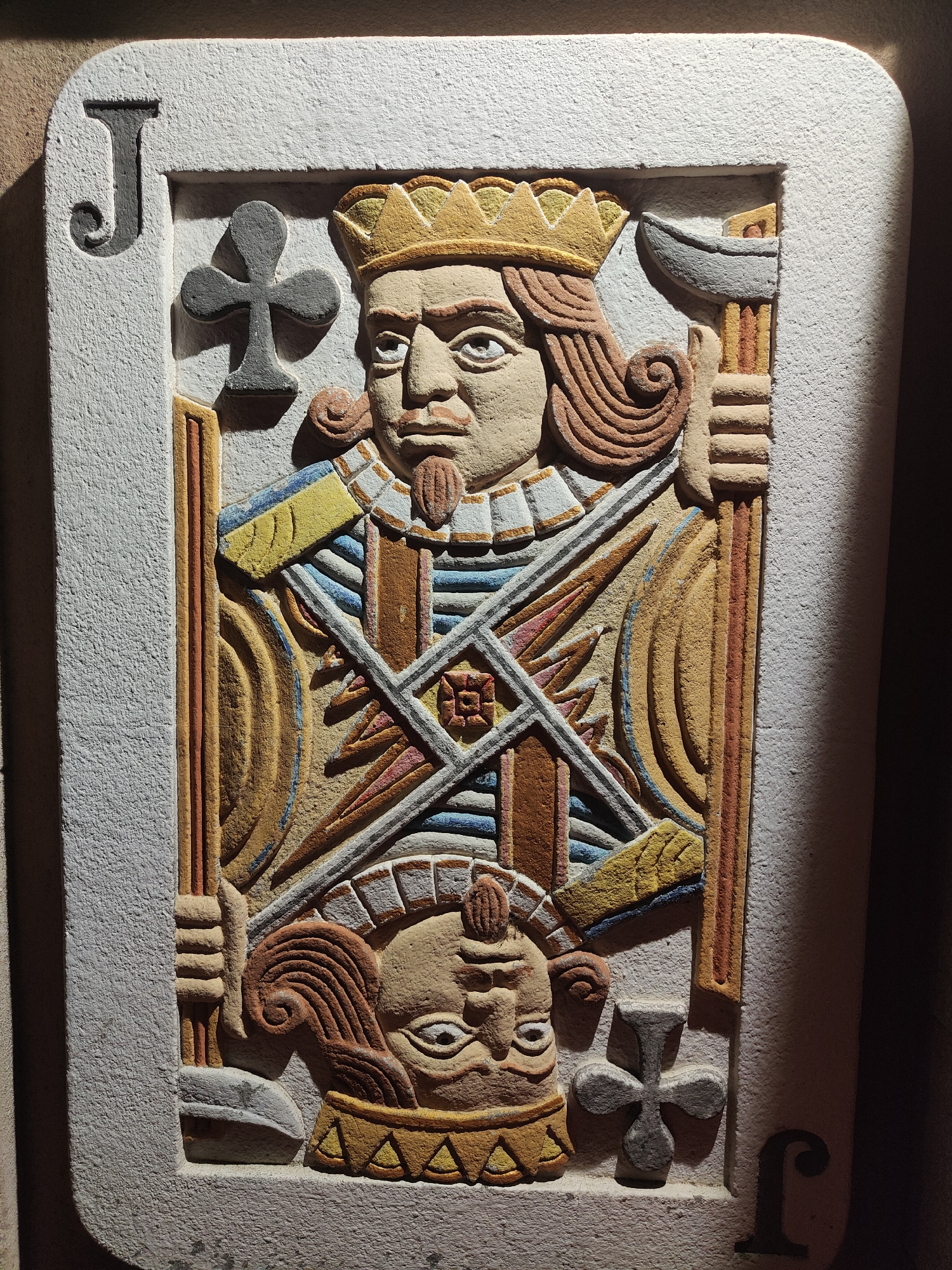A carved and painted rectangular relief, resembling a playing card, is mounted vertically on a textured, light brown wall in Burgas, Bulgaria. The relief measures approximately 60-70 cm in height and 35-40 cm in width, featuring rounded corners and a raised, off-white border. The surface texture is rough and granular, indicative of concrete or coarse plaster.
The central design depicts a stylized, full-length figure consistent with a Jack of Clubs. In the upper left and lower right corners of the border, a dark grey capital "J" with a decorative serif is present. Adjacent to each "J", a dark grey club symbol (♣) is embedded in the relief.
The central figure is rendered in a traditional playing card style, with the top half showing the main depiction and the bottom half an inverted, truncated representation of the same figure. The main figure features a pale, beige-colored face with dark, wide eyes, a prominent nose, and a small, pointed dark brown goatee. A yellow-gold crown with darker accents rests on wavy, reddish-brown hair that curls at the sides. The figure wears a multi-colored garment featuring a patterned collar, a blue and white striped tunic, and elements of brown and ochre armor or clothing. The figure's hands, also beige-colored, grip two distinct objects. The figure's left hand (viewer's right) holds an upright, long, thin, brown staff or sword, partially obscured by the border. The figure's right hand (viewer's left) firmly grasps a larger, textured staff topped with a grey, crescent-shaped axe blade or halberd head.
The inverted lower portion of the relief displays the top of the same figure's head and shoulders. This includes an inverted yellow-gold crown, an upside-down beige face with visible eyes and facial features, and wavy, reddish-brown hair.
The entire relief is illuminated by a strong, directional light source from the upper right, casting pronounced shadows towards the left and downwards, accentuating the three-dimensional quality and coarse texture of the carving.