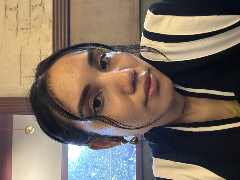 A headshot-style image features a young adult female, appearing to be of South Asian descent, positioned indoors within a location in Kolkata, India. She has a medium complexion, dark hair styled partially pulled back with some strands framing her face, dark eyes, and full lips. She is wearing a black jacket with prominent white vertical stripes on the sleeves and collar, with a yellow garment visible underneath. A healed scar is visible on the left side of her neck. A gold-colored, bell-shaped drop earring is worn in her left ear. The subject directly faces the viewer. The background on the left features a window revealing blurred green foliage outside. To the right and behind her is a light-colored, textured wall with a subtle brick-like pattern, bordered by dark wooden trim. A small, spherical, gold-colored decorative object hangs from the ceiling in the upper left background. The setting suggests an indoor public space, possibly a cafe or restaurant, illuminated by natural light from the window.
